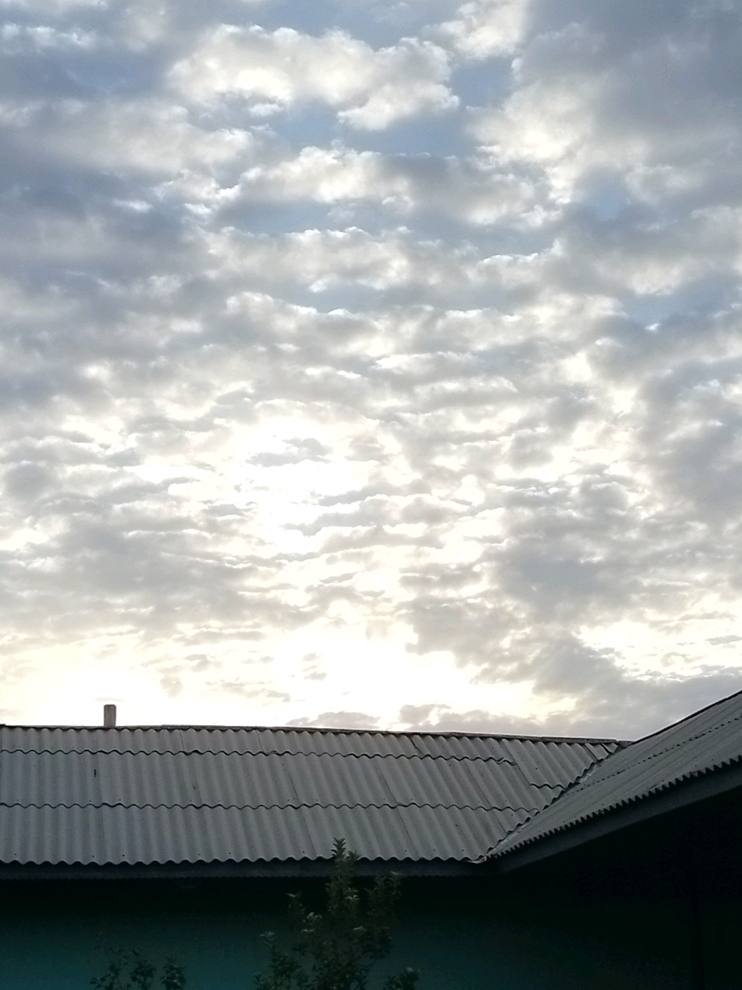 The image shows a cloudy sky over the roofs of several buildings. The clouds are a mix of white and gray, with some patches of blue sky visible. The roofs are made of corrugated metal and appear to be weathered. There is a chimney visible on one of the roofs. Some trees are visible in the foreground. The image is taken from a low angle, looking up at the sky. The location is identified as Kölkent, Kazakhstan.