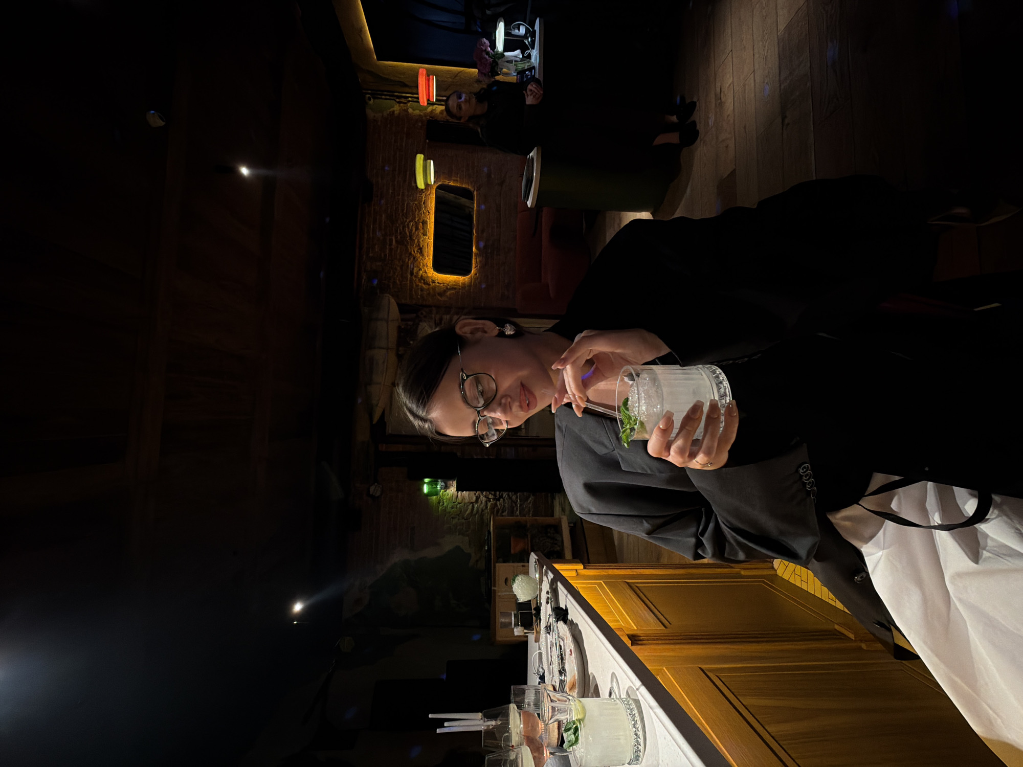 In a dimly lit interior, possibly a bar or restaurant, a woman with glasses and dark hair is seated at a counter. She is wearing a black blazer over a black garment and is holding a clear glass with a light-colored beverage and ice, garnished with mint. Her left hand is raised, and she appears to be touching the straw in the glass. The counter is made of a light-colored material with a wooden base, and several other glasses are visible on it, along with some plates and cutlery. In the background, exposed brick walls are visible, and colorful pendant lights hang from the ceiling. Another woman, wearing a dark dress, stands behind the counter in the mid-ground. The floor is made of wooden planks. The image is taken at eye level, with the focus on the seated woman and her drink. The scene appears to be a social setting, likely in Krasnoyarsk, Russia.