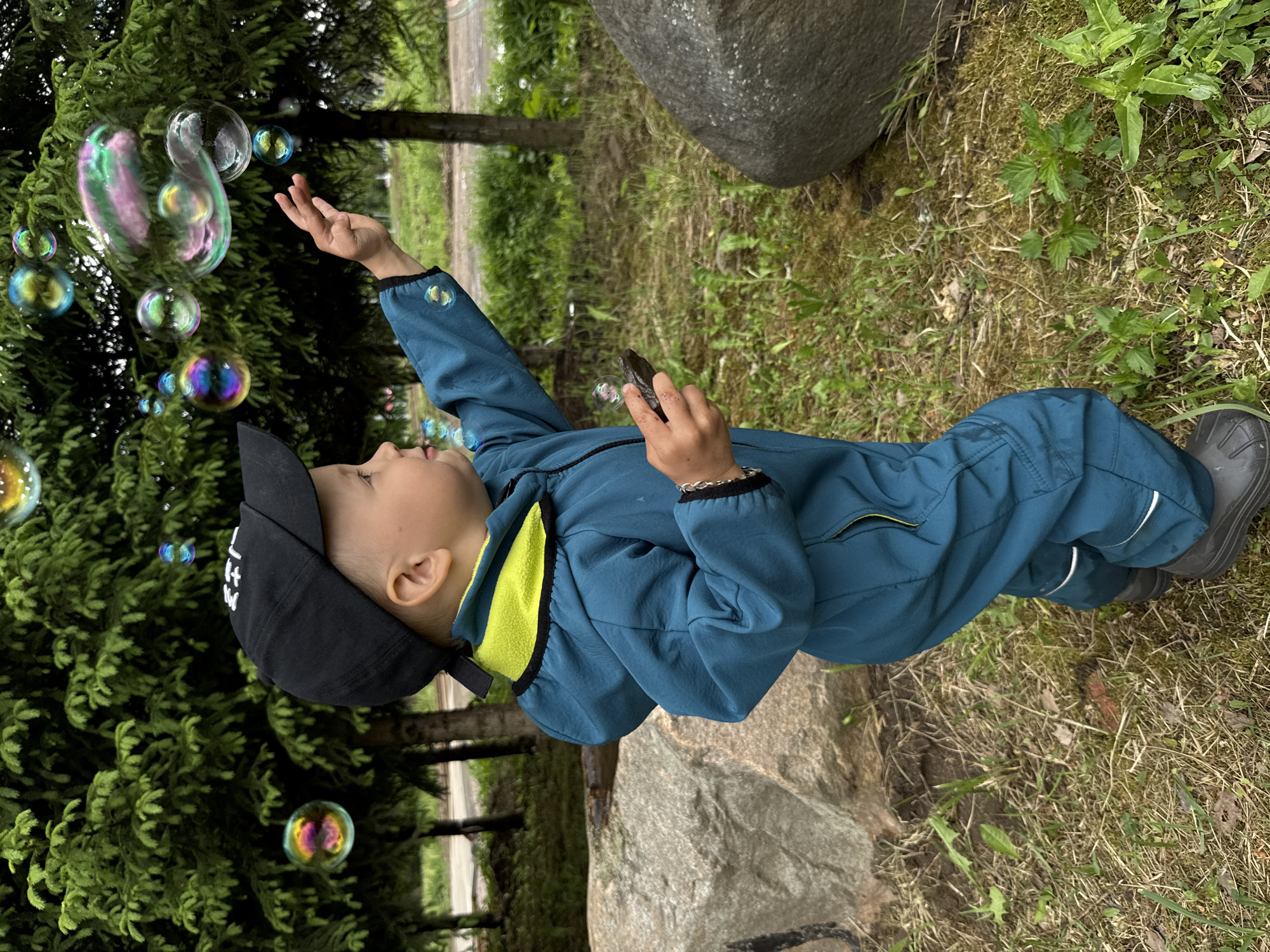 A young child wearing a dark-colored baseball cap and a blue jumpsuit with yellow accents is reaching up towards soap bubbles. The child is outside, with a large evergreen tree to the left. The ground is covered with grass and other vegetation, and there is a large stone or boulder nearby. Based on the location provided, this scene is likely taking place in the Rusanovo district of Russia.