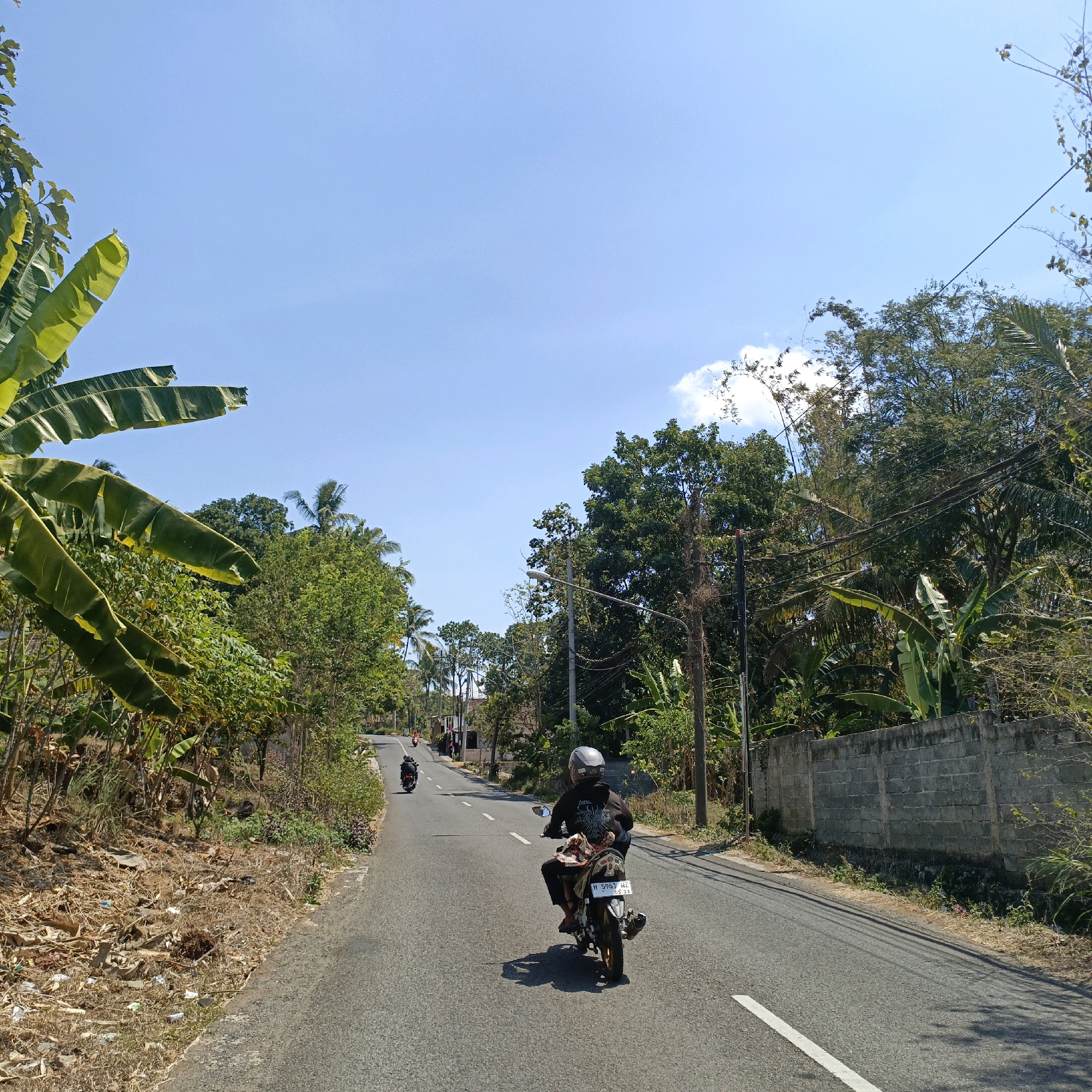 This is a wide, eye-level shot capturing a paved road in Krajan Lor, Indonesia, on a bright, clear day. The road has a white dashed line down the center and a solid white line on the right side. A person wearing a helmet and a dark hooded jacket is riding a motorcycle forward on the road. The motorcycle has a license plate that reads "H 5961 HZ 08-23". In the distance, another motorcycle with two riders is visible.
The environment is lush and tropical. On the left side of the road, there are tall banana trees with large leaves, dry brush, and scattered debris. On the right side, a grey cinder block wall runs along the road, behind which are dense trees and foliage, including more banana trees and palm trees. Electrical poles and wires are present along the roadside, with one pole situated prominently in the foreground on the right. The sky is a clear, bright blue with a few scattered white clouds. The sunlight casts shadows on the road, indicating the time of day is likely late morning or early afternoon. The overall impression is a rural or semi-rural tropical setting.