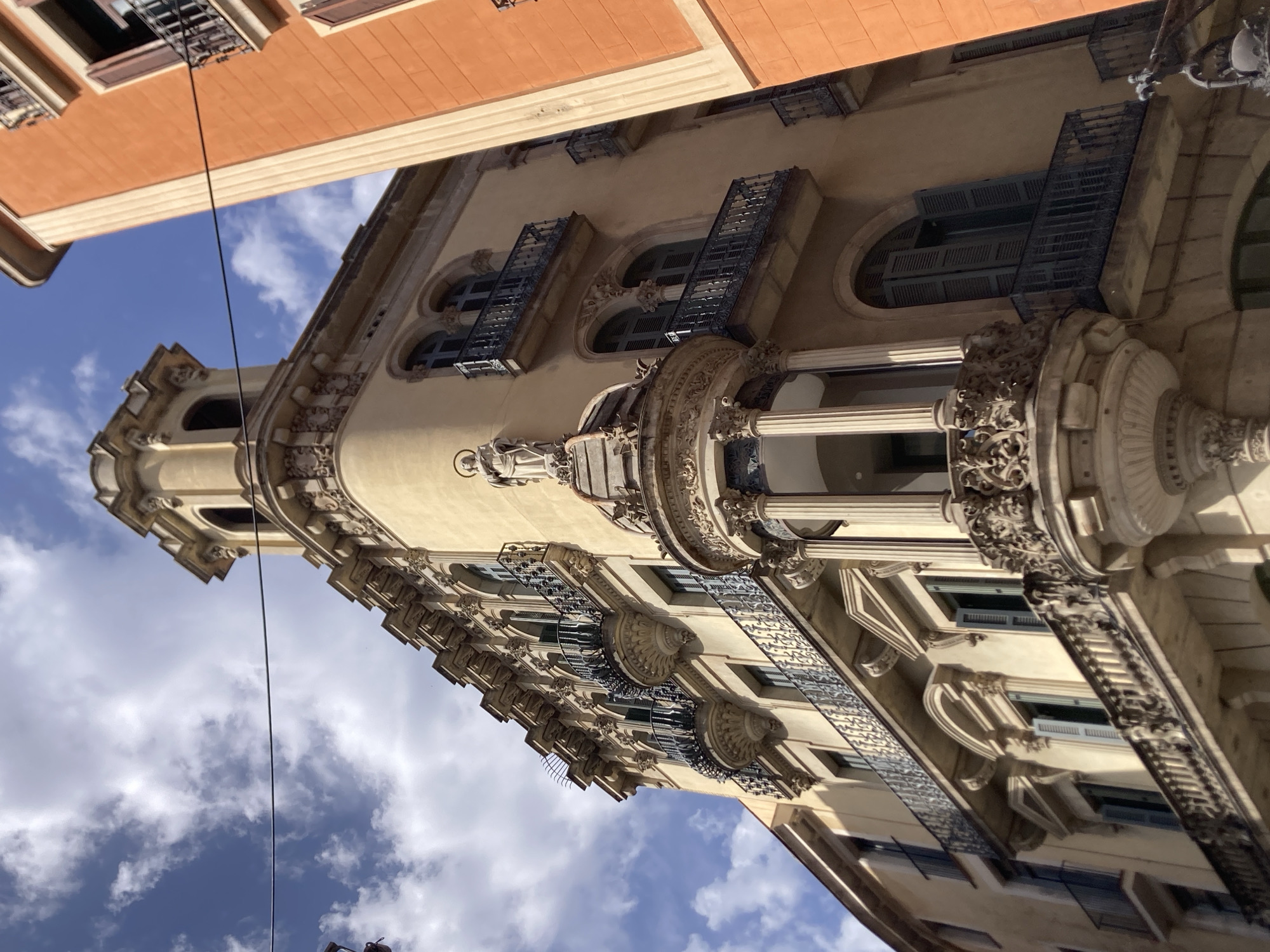 The image shows a close-up, low-angle view of the facade of a historic building in Barcelona, Spain. The building is light yellow and features ornate architectural details, including balconies with intricate iron railings, decorative cornices, and arched windows with green shutters. A statue of a robed figure with a halo stands atop a rounded structure on the building's corner. The building is adjacent to a bright orange building on the right side of the frame. The sky above is blue with scattered white clouds. The perspective suggests the photo was taken from a street level looking upwards.