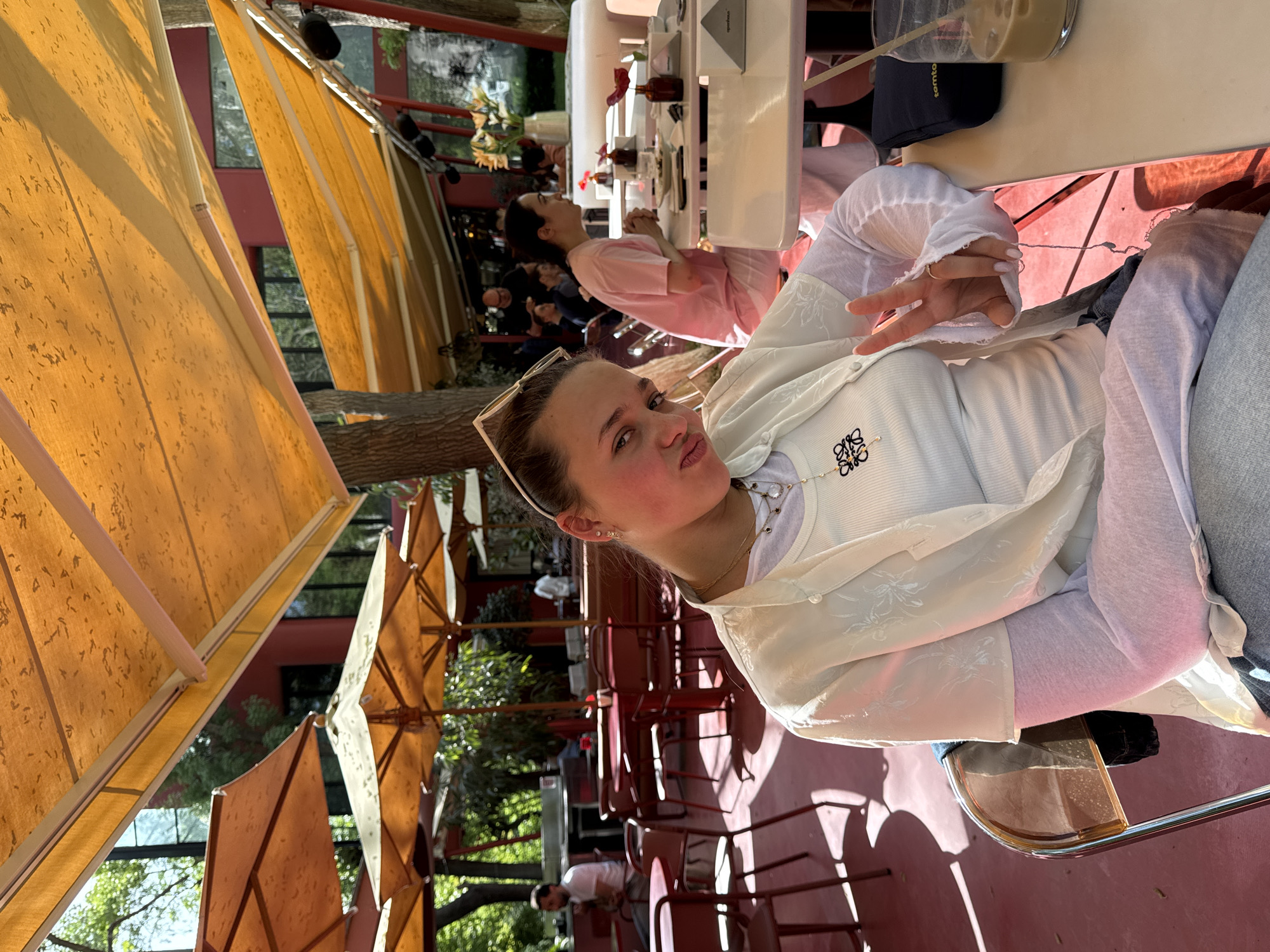 The image shows a young woman sitting at an outdoor cafe, likely in Yerevan, Armenia, though this location is not directly verifiable from visual elements. She's fair-skinned with light brown hair and is wearing sunglasses perched on her head. She has on a white cardigan over a white top with a logo and a necklace. She is making a "peace" sign with her fingers, looking towards the camera.
Behind her, the cafe setting is noticeable: yellow awnings provide shade over red chairs and tables. Other patrons are seated at tables, one wearing a pink shirt. The ground is red.