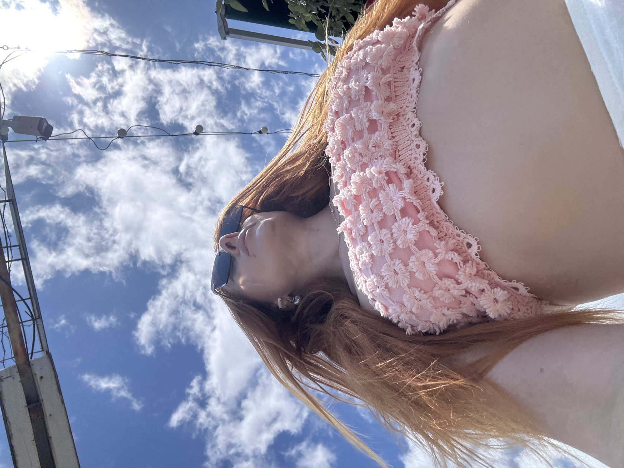 A low-angle, upward-looking shot captures a person with long, flowing reddish-brown hair against a bright blue sky. The individual, seen from the chest up and turned slightly to the right, wears dark sunglasses and has their head tilted back, looking upwards. A small, pearl-like earring is visible on their left ear. They are dressed in a light pink top featuring a delicate, possibly crocheted or embroidered, floral pattern, with a white garment visible on the right side of the frame.
The background is dominated by a vibrant blue sky dotted with scattered white cumulus and cirrus clouds. A brilliant sun in the upper left corner casts a strong light, creating noticeable lens flare. Overhead, several dark electrical wires crisscross the frame, with small light fixtures or bulbs suspended from them. To the far left, a tall, metallic structure, possibly a pole or part of a building, extends upwards. The scene depicts a clear, sunny day, suggesting warm and pleasant weather. There is no visible text in the image. The setting is an outdoor environment in Budva, Montenegro.