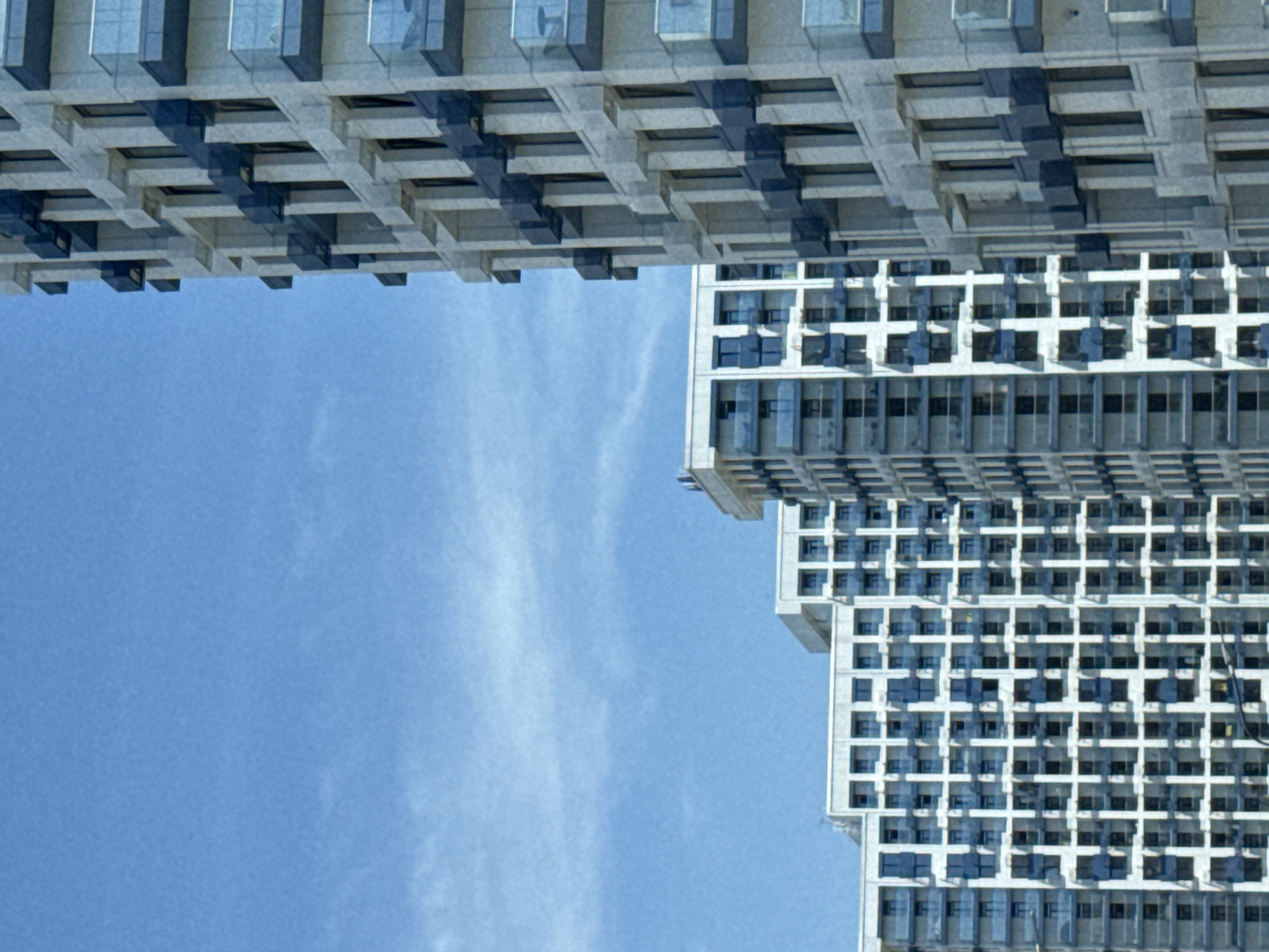 This image depicts a cluster of modern high-rise residential buildings under a clear blue sky in Sochi, Russia. In the foreground, positioned on the right side, is a tall, light-grey building with a textured facade. This building features numerous dark blue/grey rectangular projections, likely balconies or decorative elements, some of which are equipped with transparent glass railings. Dark-tinted windows are recessed into the facade. Extending from the midground to the left, several similar contemporary buildings are visible, characterized by white or light-grey exteriors and a prominent grid pattern of dark-framed windows and dark blue/grey balcony structures. The sky above is predominantly clear blue, with minimal, faint white cloud wisps visible in the lower-left quadrant. No individuals or specific actions are discernible within the frame. The scene suggests a modern urban residential environment.