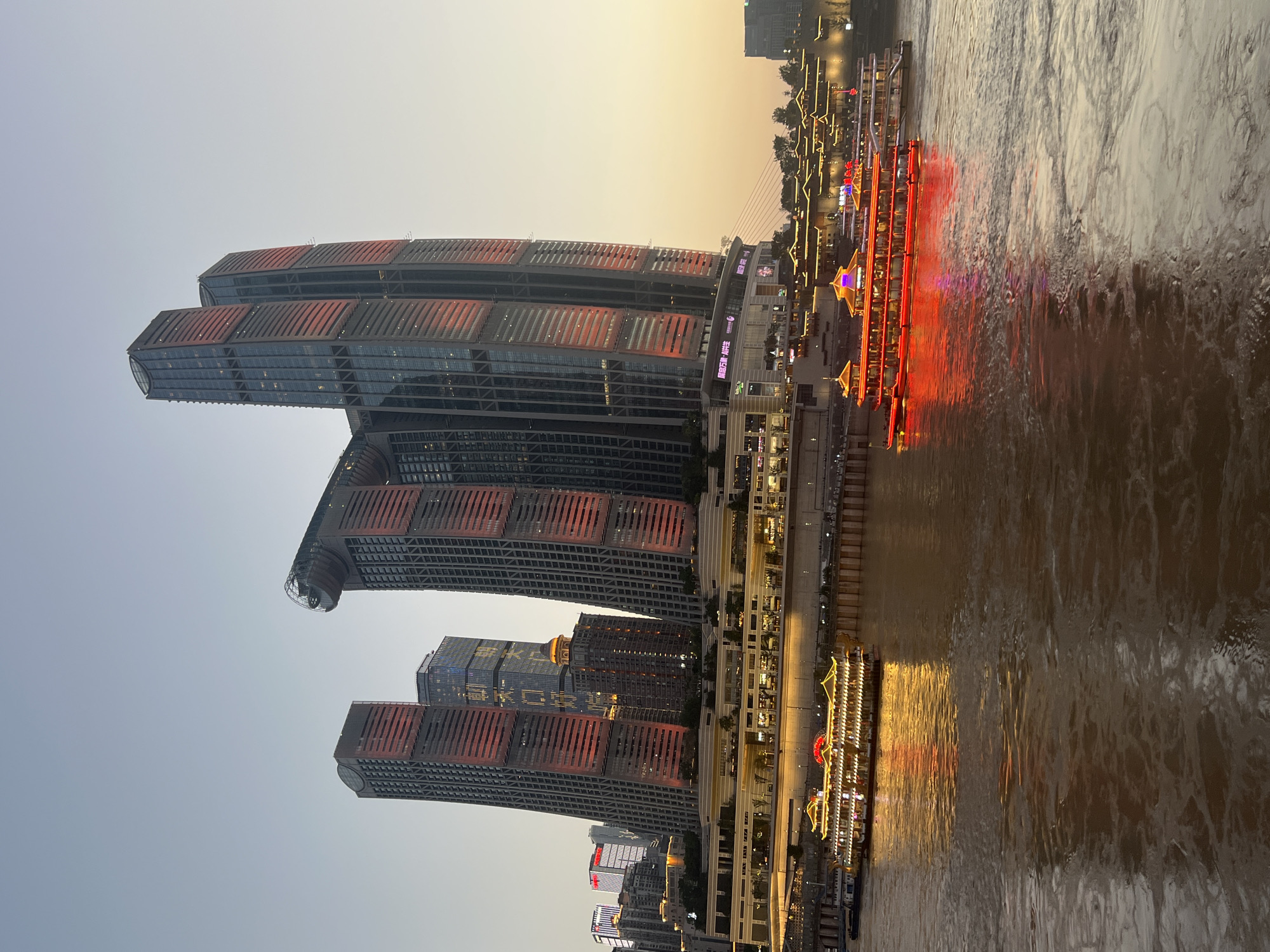 The image depicts the Raffles City Chongqing complex, a distinctive landmark located in the Nan'an District of Chongqing, China. The complex features several towering skyscrapers, with the most prominent being the twin towers that form an iconic "sail" shape. The buildings are illuminated with red and orange lights, giving them a vibrant glow against the dusky sky. Below the skyscrapers, a multi-level waterfront promenade is visible, lined with illuminated buildings and trees.
In the foreground, a wide river flows, reflecting the lights of the city and the vessels on the water. Two passenger boats are prominently featured. The boat on the left is decorated with traditional Chinese architectural elements and illuminated with yellow lights. The boat on the right is a modern vessel, aglow with bright red lights. The water appears somewhat murky, suggesting it is a substantial river. The overall atmosphere is one of a bustling, modern city at dusk, with the lights of the buildings and boats creating a picturesque scene. The sky is a gradient of pale blue and warm orange, indicative of sunset or twilight.