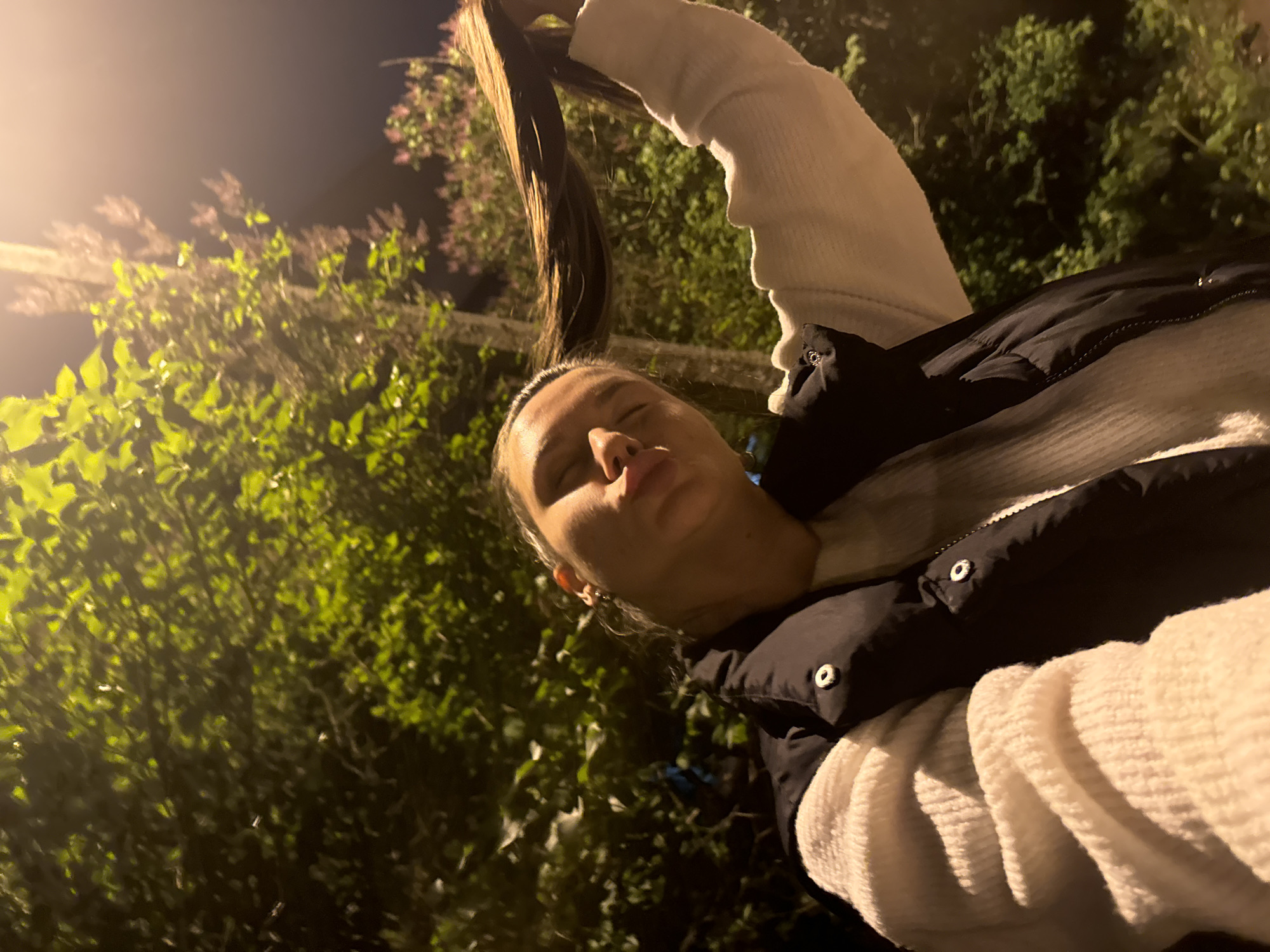 A young woman is posing for a photo in Vniissok, Russia. She is holding her hair up in her right hand, and her face is angled towards the camera. The woman is wearing a white sweater, and a black puffer vest. The background includes greenery and a street light.
