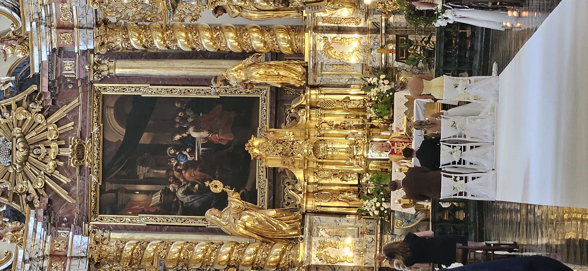 The image shows a wedding ceremony taking place inside a richly decorated church, likely St. Mary's Basilica, located in Krakow, Poland. The altar area is adorned with ornate gold elements, including statues of religious figures and intricate carvings. A large painting depicting a biblical scene hangs above the altar. In the foreground, a wedding party is seated in front of the altar. A bride in a white wedding dress is seated next to a groom in a brown suit. Other guests are visible, including a woman in a black dress on the left. A priest in religious vestments stands at the altar, conducting the ceremony. The church interior features marble columns, gilded decorations, and a white carpet runner leading to the altar. Candles and floral arrangements enhance the celebratory atmosphere.