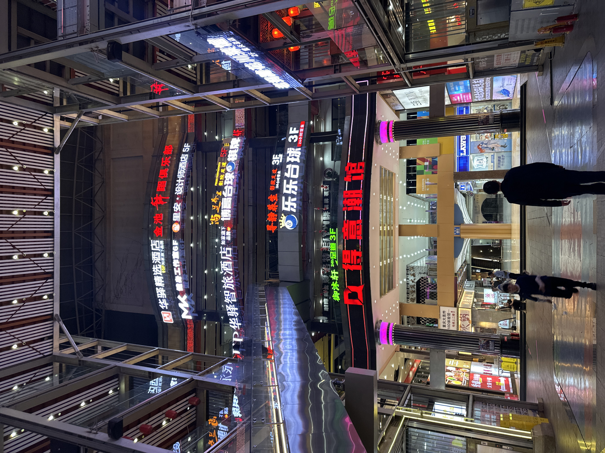The image shows a night view of a multi-level commercial area in Yuzhong District, China. Various businesses are advertised with neon signs in Chinese characters. People are walking along a polished floor. A steel and glass structure is visible above. The signs indicate establishments such as hotels, gyms, design studios, billiard rooms, and restaurants. Some establishments also have signs in English.