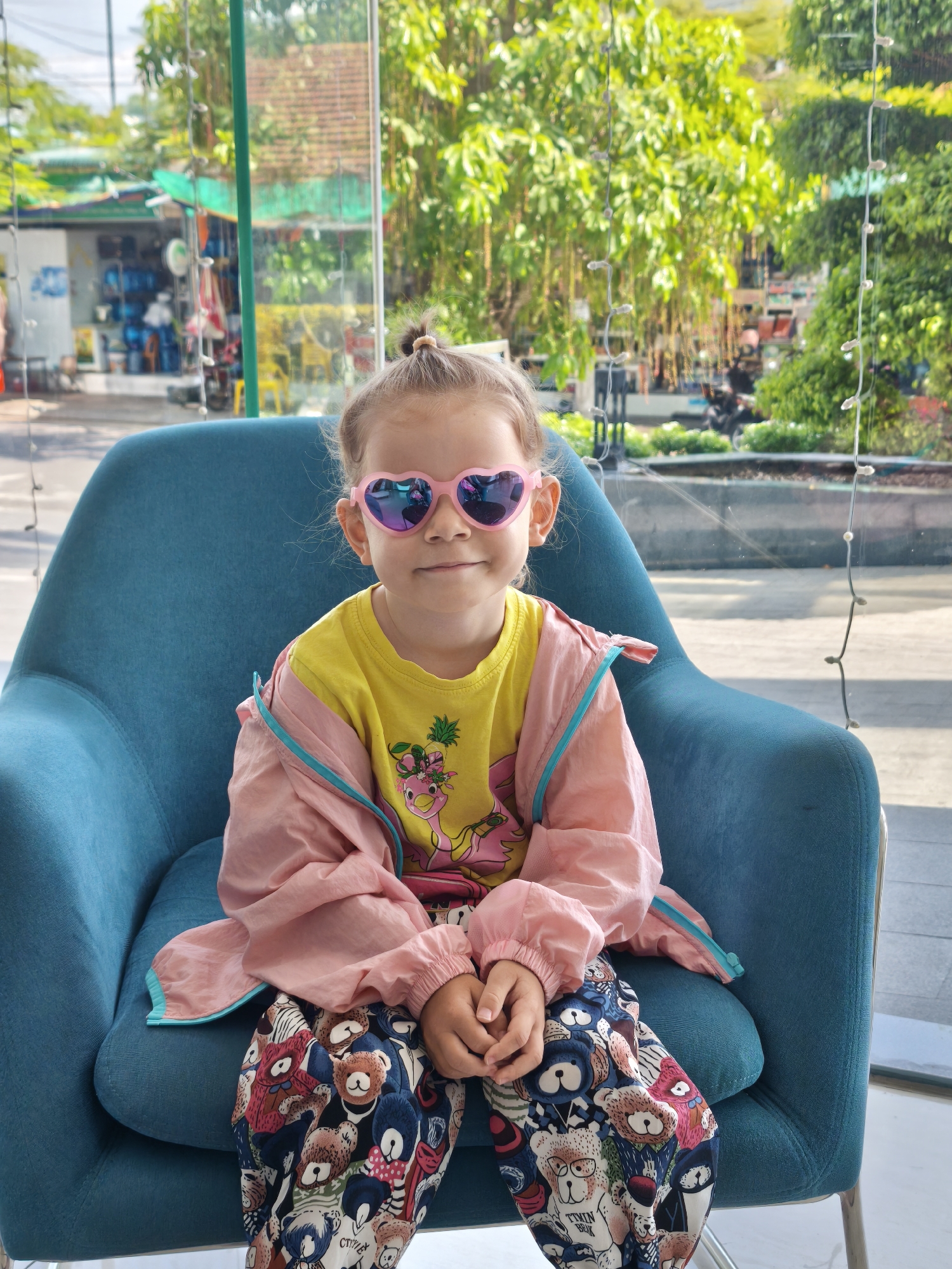 A young girl with blonde hair styled in a top knot and wearing heart-shaped pink sunglasses sits in a teal armchair. She has on a yellow t-shirt with a cartoon flamingo graphic and a pink and turquoise jacket. Her pants are printed with a pattern of teddy bears. The girl is facing the camera and smiling. She is seated indoors, with a large window behind her that looks out onto a street scene with trees and buildings. The location appears to be Bac Nha Trang Ward, Vietnam.