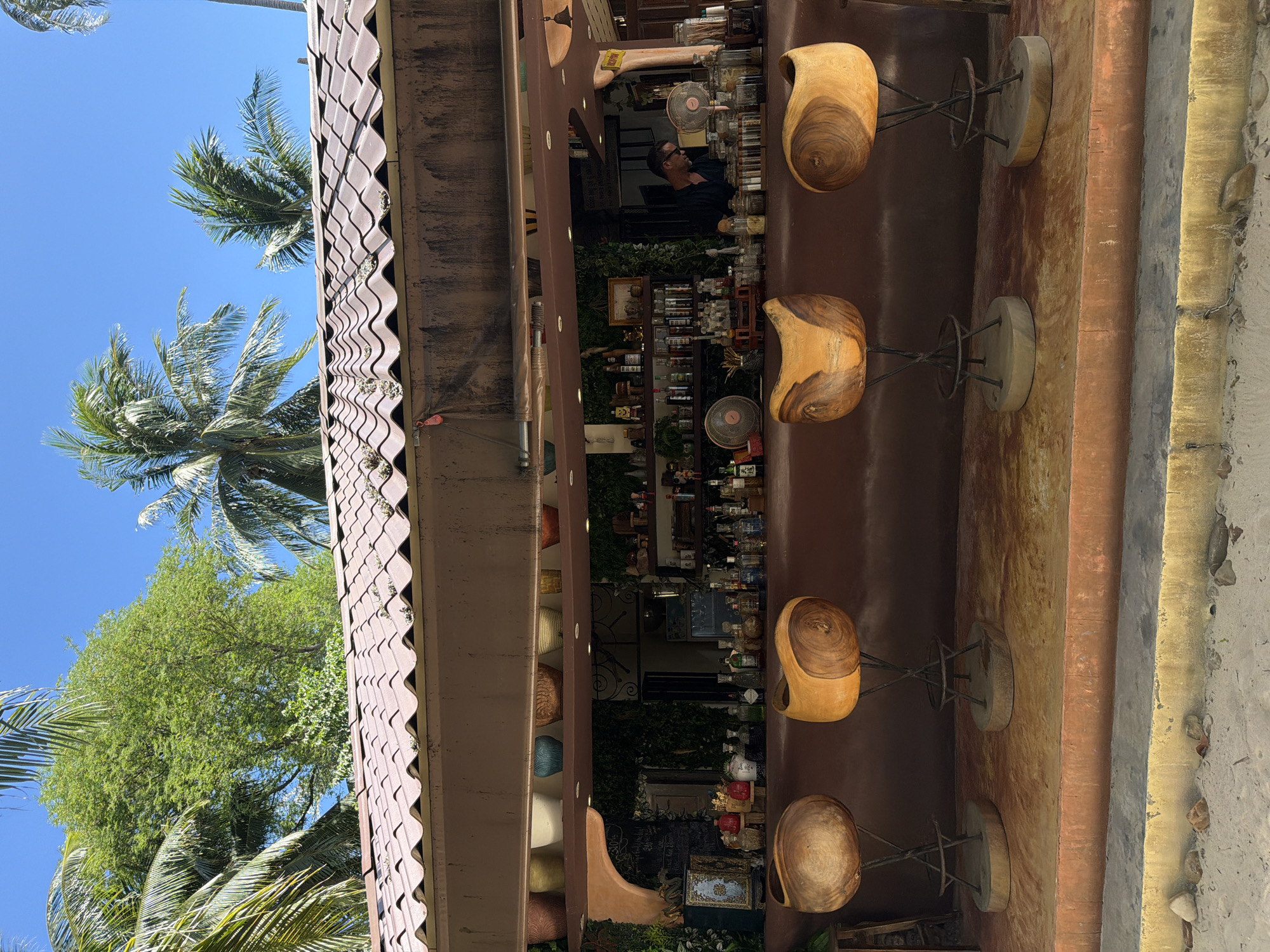 This image depicts an outdoor bar area with a covered roof in Baan Thong Krut, Thailand. Four unique wooden bar stools with metal legs are arranged in front of a long, curved bar. Behind the bar, shelves are stocked with various bottles, jars, and decorative items, including what appear to be small fans. A man wearing glasses and a dark shirt is visible behind the bar, seemingly working. Lush green foliage and palm trees are visible above and behind the structure, suggesting a tropical setting. The ground in front of the bar is sandy, with some small rocks scattered about. The overall scene suggests a relaxed, open-air bar environment.