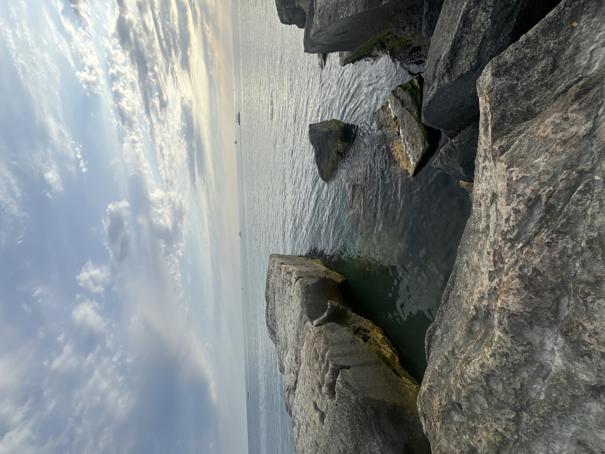 The image depicts a rocky shoreline with calm water and a cloudy sky. Large, rough-hewn gray rocks are prominent in the foreground and midground, some partially submerged in the dark water. The water appears clear enough in some areas to see the rocky bottom. In the distance, several small boats are visible on the horizon, and the sky is filled with scattered, wispy clouds tinged with the soft light of either sunrise or sunset. The scene has a natural, unpopulated feel.
This scene is consistent with the coastline of the city of Aktau, Kazakhstan, particularly areas along the Caspian Sea that feature natural rock formations and a tranquil, open water environment.
