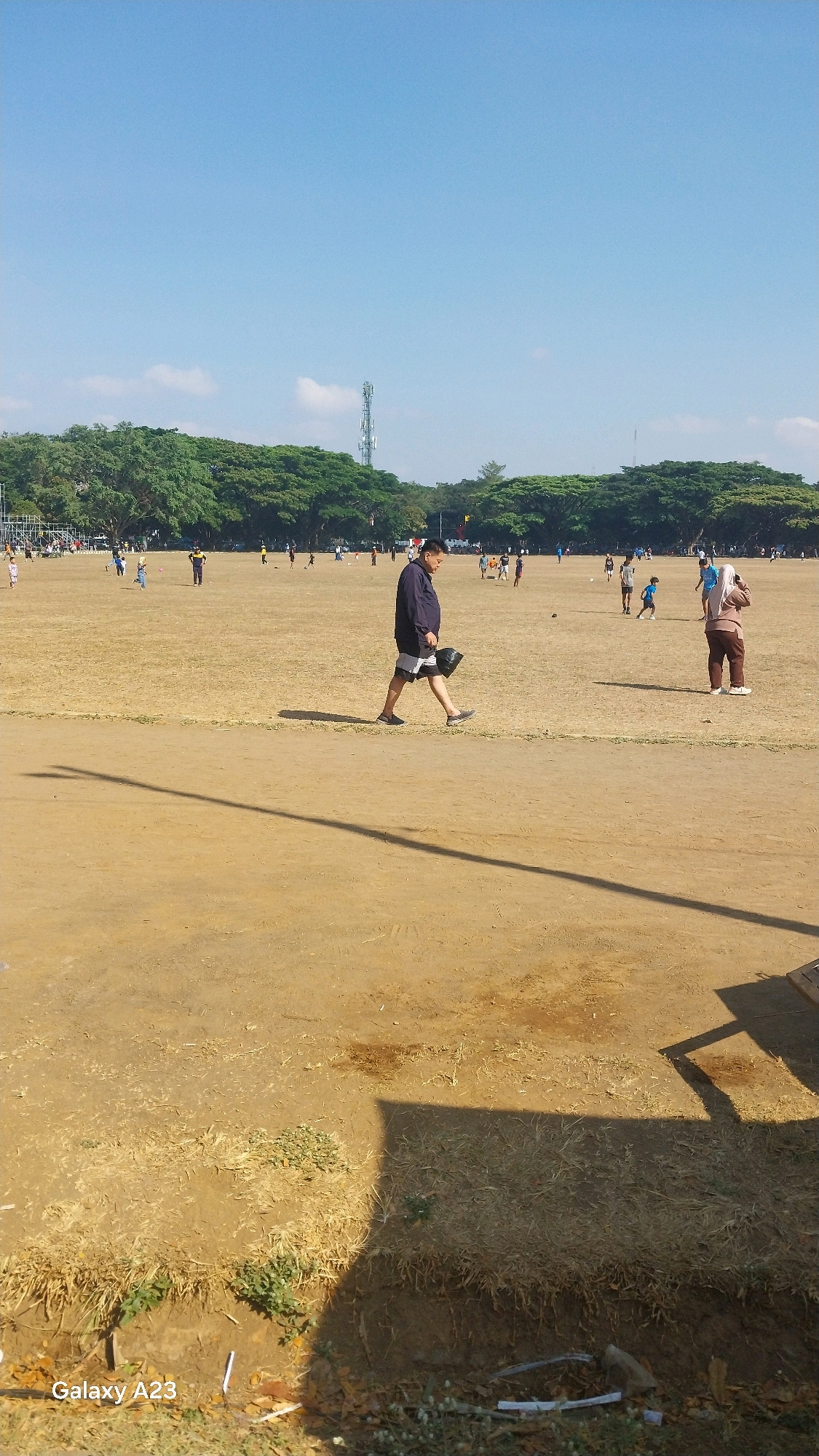 The image shows a wide, dusty field with scattered groups of people playing sports or observing. In the foreground, a man in a dark blue shirt and shorts walks to the right, carrying a black bag. Further back, other individuals are engaged in activities, some appearing to be playing soccer or a similar game. A woman in a beige hijab and brown pants stands with her hands near her face, looking off to the side. The field is surrounded by a dense line of trees, and in the background, a tall telecommunications tower is visible. The sky is clear and blue. The "Galaxy A23" watermark in the bottom left corner indicates the device used to capture the image, and while the location is not explicitly stated in the image, this scene is typical of outdoor sports fields found in the city of Malang, Indonesia.