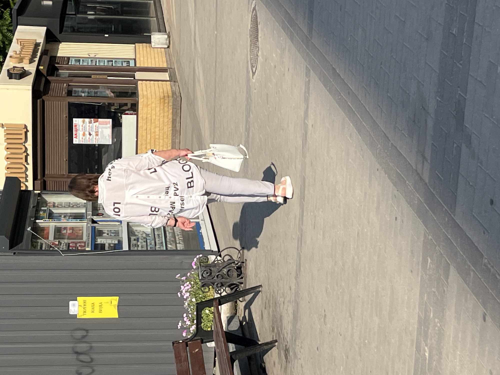 A person walks down a sunlit sidewalk in Dnipro, Ukraine. The individual is wearing a white shirt with black lettering and light-colored pants, carrying a white handbag. The shirt displays text in various sizes, including "BLOC" and "107". The setting appears to be an urban area with shops lining the left side of the street. One shop has a sign that says "АКЦІЯ!", indicating a sale or promotion. A yellow sign with Cyrillic text is attached to a grey wall on the left. A wooden bench with a wrought-iron planter filled with pink flowers sits on the right side of the sidewalk. The lighting suggests it is daytime, likely mid-morning or afternoon, with clear weather as indicated by the distinct shadow cast by the person. The overall scene depicts a casual stroll through a commercial district.