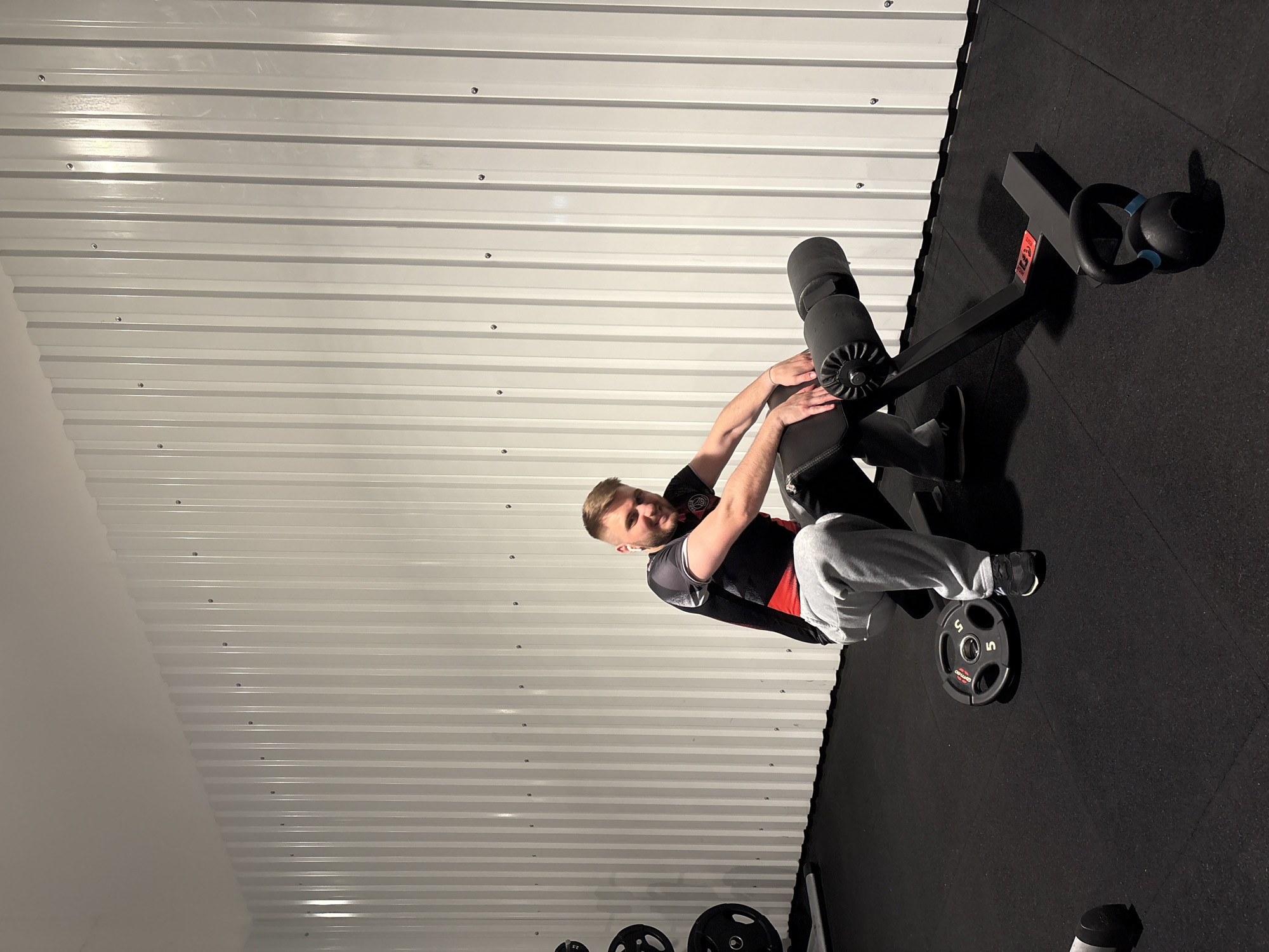 A man with short blond hair and a beard, wearing a black and red athletic shirt and grey sweatpants, sits on a black exercise bench in a gym. He is smiling and looking towards the camera. His hands are resting on the bench. To his left, on the black rubber floor, is a 5-kilogram weight plate. To his right, also on the floor, is a black kettlebell with blue accents. The background features a white, corrugated metal wall with visible screws. On the far left of the image, a rack with weight plates is partially visible. The gym appears to be located in Mönchengladbach, Germany.