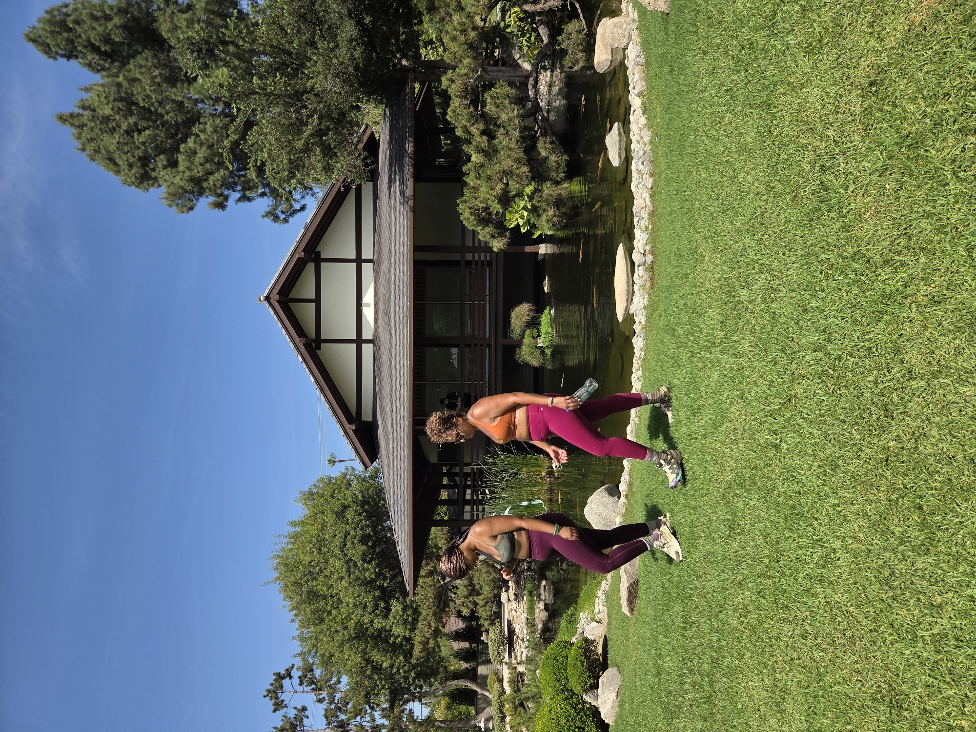 The image depicts a serene outdoor scene with two women walking on a lush green lawn in front of a Japanese-style building. The building features a traditional dark-tiled roof with exposed wooden beams and white walls. A tranquil pond with koi fish is visible in front of the building, bordered by rocks and lush greenery. The woman on the left, wearing a green sports bra and dark purple leggings, is looking down at her phone. The woman on the right, in an orange sports bra and bright pink leggings, is holding a water bottle and looking ahead. Both women are wearing athletic shoes. The background consists of mature trees and a clear blue sky. The setting appears to be a botanical garden or a cultural center in Glendale, California.