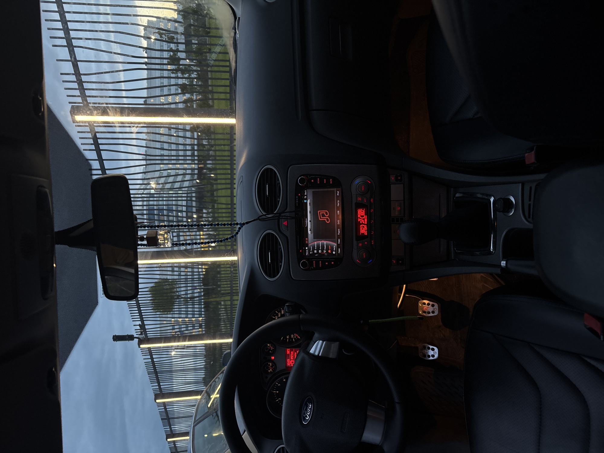 The image shows the interior of a Ford Focus car, likely parked, looking out through the windshield. The time on the dashboard indicates 13:15. The car is situated in a location that features a distinctive metal fence with vertical LED lighting. Beyond the fence, there are trees and a modern building with many windows, suggesting an urban or semi-urban environment. The sky is overcast. The driver's seat, steering wheel, dashboard with a Ford logo, car stereo, and gear shifter are visible. The pedals are also in view. The overall lighting suggests it is daytime, possibly late afternoon or early morning, given the overcast conditions and the artificial lights being on. The location is identified as Arfa, Russia.