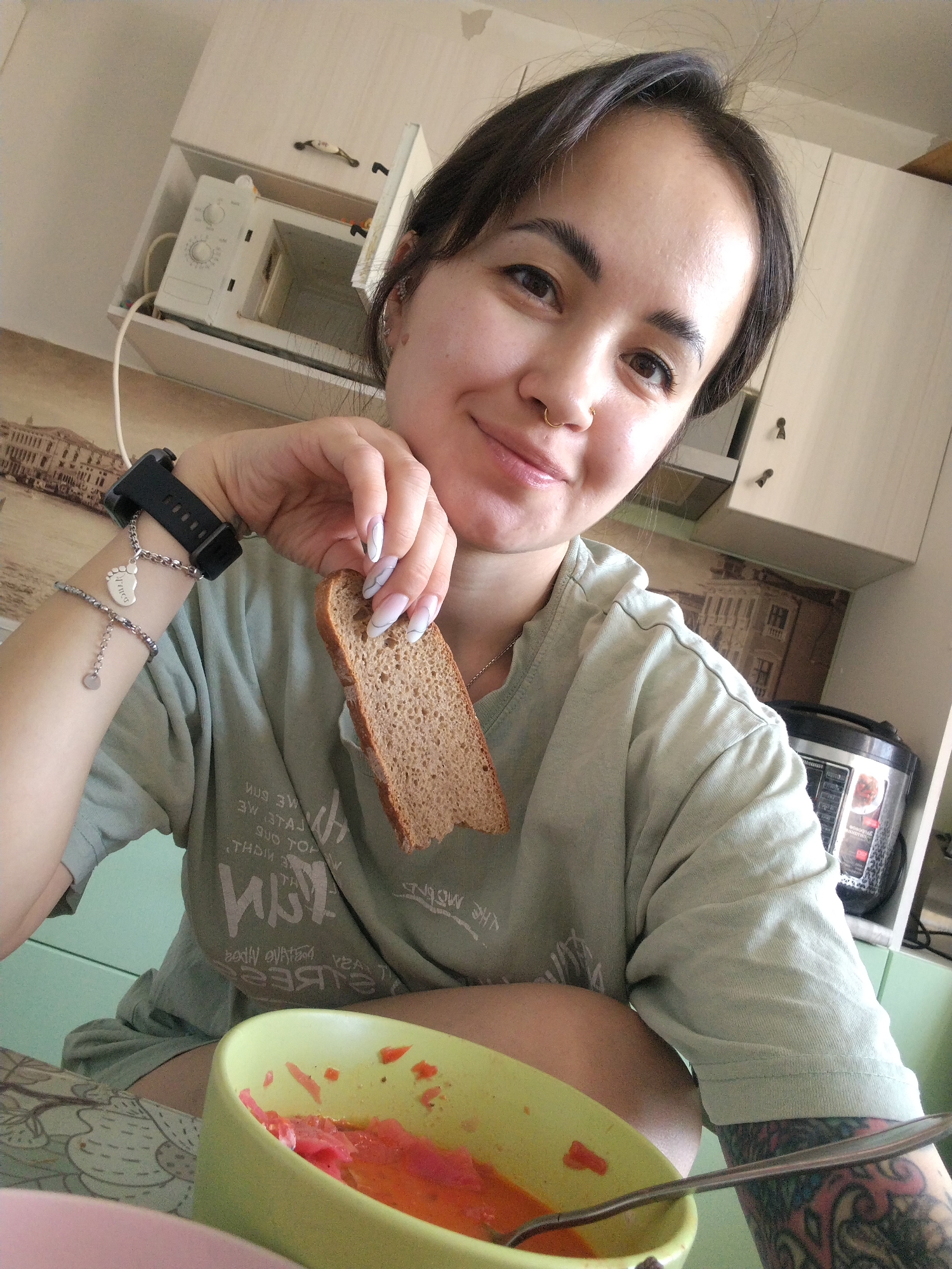 A woman is in a kitchen, eating. She is holding a piece of brown bread in her right hand. She has a bowl of food in front of her, which she is eating with a spoon. She has a watch and a bracelet on her left wrist. There is a microwave in a cabinet above her. A multi-cooker is on the countertop behind her. A building mural is on the backsplash above the kitchen counter. According to reverse image search, the building mural is a depiction of a historical building in Noviy, Russia.
