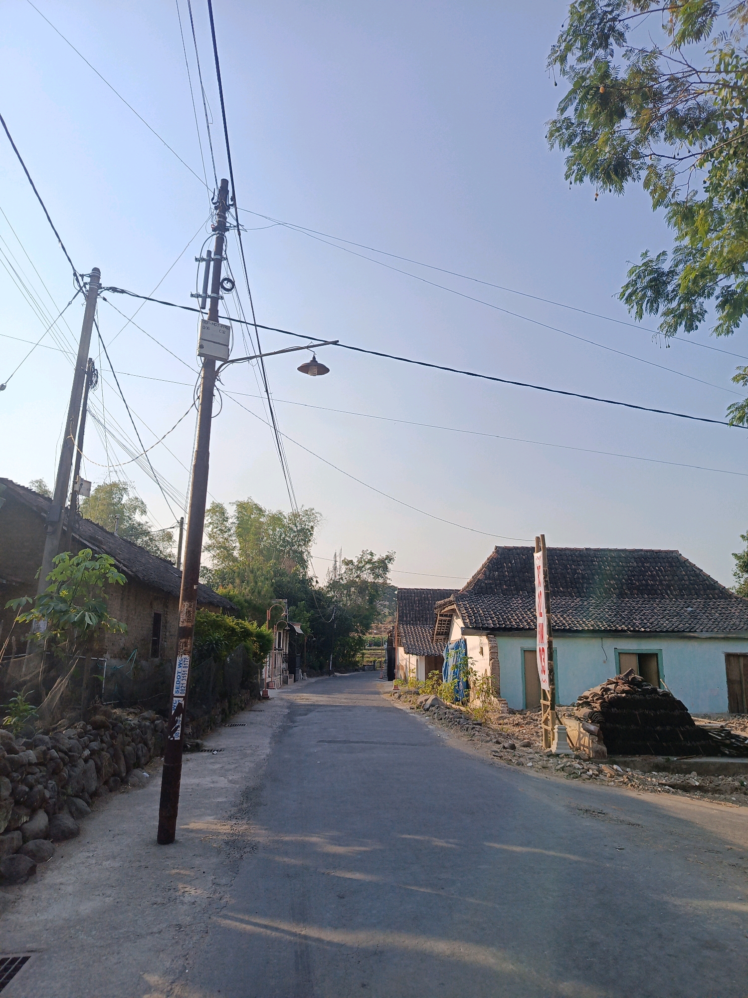 The image depicts a quiet, sunlit street in Ringinagung, Indonesia, with traditional Indonesian houses lining the road. The sky is clear and blue, with a few scattered tree branches visible in the upper right corner. A prominent utility pole with numerous wires stands to the left of the road, adorned with a small junction box and a street lamp.
The houses are single-story structures with tiled roofs, common in rural Indonesian architecture. The house on the right has a light blue facade and is surrounded by a pile of dark tiles, suggesting recent construction or renovation. The house on the left appears older, with a rough, textured exterior wall and a stone foundation.
The road itself is paved and appears to be in good condition, with shadows cast by the utility pole and trees across its surface. The area is sparsely populated, with no people visible in the frame. The overall impression is one of a peaceful, rural setting bathed in the warm light of the sun.