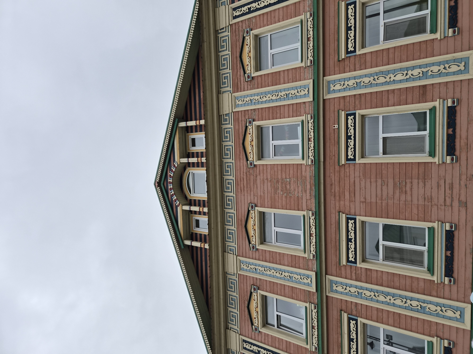 This is a low-angle, eye-level exterior shot of a historic wooden building with ornate architectural details, likely located in Kazan, Russia. The building's facade is primarily pinkish-brown wood siding with multiple rows of windows. The windows are modern, with white frames and clear glass.
The architectural embellishments are a prominent feature. There are decorative wooden elements around each window, painted in yellow, green, and blue. Above the second row of windows, a Greek key-like pattern is visible in blue and yellow. The top of the building features a gabled roof with striped siding in brown and blue, and intricate decorative trim along the eaves. Several small dormer windows are visible on the upper part of the roof. The sky above is overcast and light gray. There are no people or animals visible in the image.