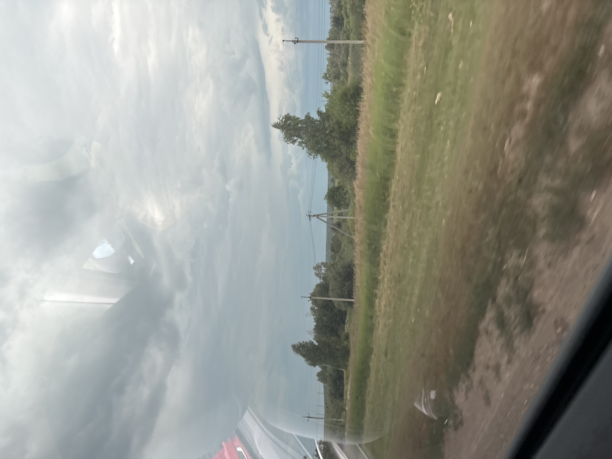 The image depicts a rural landscape seen through a window, likely from a moving vehicle. The foreground shows a dirt and grassy roadside. Beyond this, a field of tall grass stretches towards a treeline. Several utility poles with power lines are visible, interspersed with trees and bushes. The sky is overcast with dramatic, dark clouds, suggesting a stormy or approaching storm. The location is identified as Krasnaya Gorka, Russia. Reflections are visible on the window, including what appears to be part of the vehicle's interior and a side mirror.
