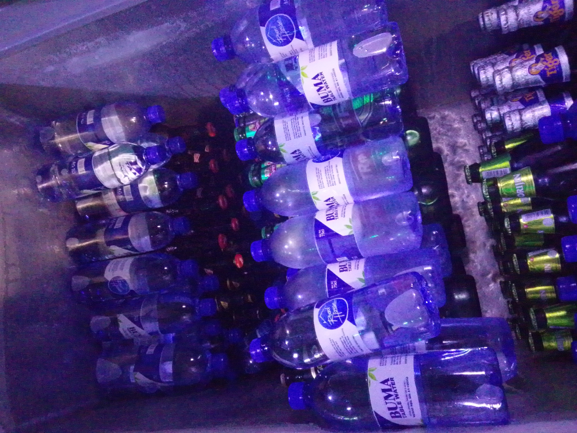 The image depicts a collection of bottled beverages, primarily water, stored in what appears to be a cooler or storage bin. The bottles are predominantly made of clear plastic with blue caps. Labels visible on some bottles indicate "BUMA TABLE WATER". In the background, a row of bottles with yellow and green labels, possibly beer, is also present. The lighting is dim and casts a purple hue over the scene, suggesting an indoor, possibly commercial or storage environment. No people or specific location markers are identifiable within the image itself, beyond the presence of these beverage containers. There is no information in the image to suggest a connection to Howayi, Nigeria.