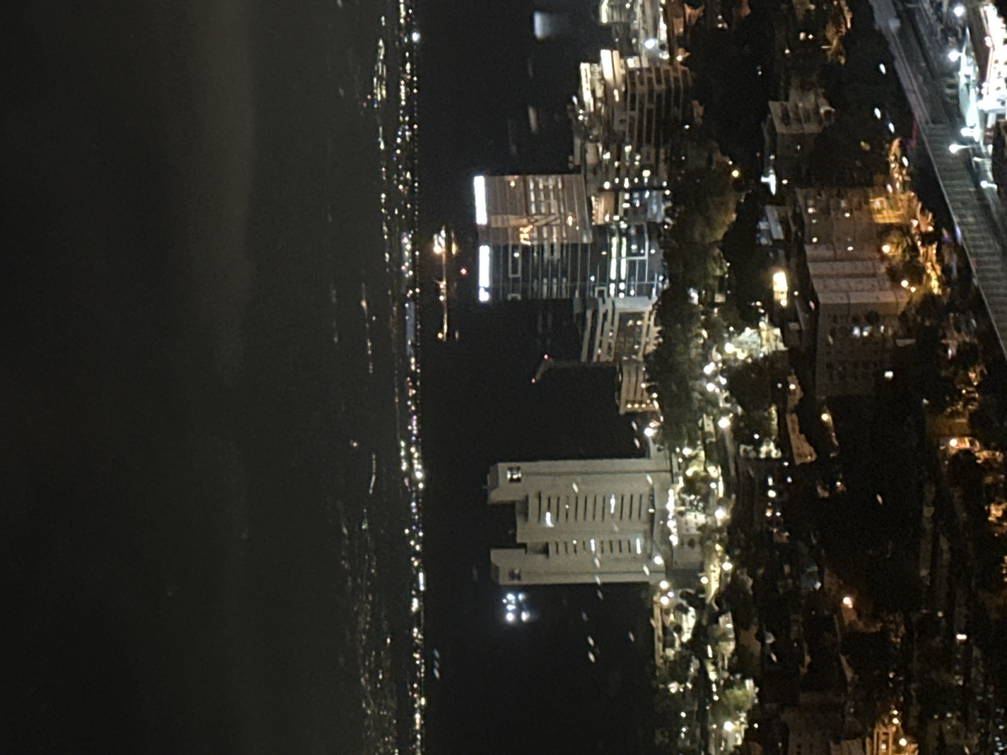 A long shot aerial photograph captures a nighttime cityscape in Haifa, Israel, featuring a body of water and several illuminated buildings. The dark sky is filled with indistinct clouds, and the distant horizon displays a blurred line of city lights. In the mid-ground, a large, light-colored rectangular building with several floors and rows of windows stands prominently. To its right, a modern, darker skyscraper with glass-like facades and bright window lights is visible. The foreground is densely populated with smaller buildings, some with illuminated windows, and scattered trees and greenery. The overall scene is one of a vibrant urban environment at night, with the water reflecting some of the artificial lights.