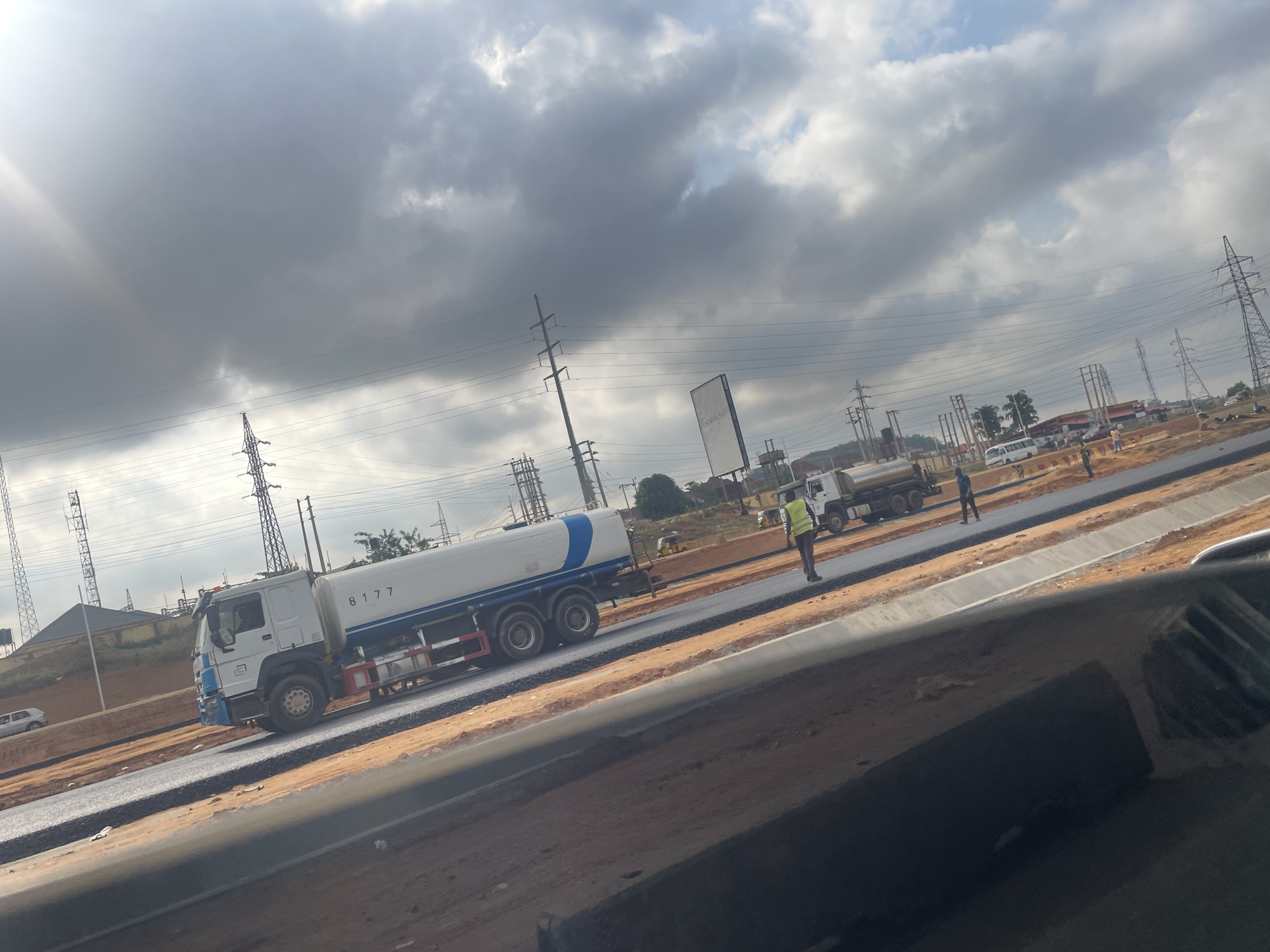 The image depicts a road construction site in Abuja, Nigeria. Two large white water tankers are positioned on a newly paved section of the road. Several construction workers are present, including one wearing a yellow safety vest. Overhead electrical power lines are visible. The sky is overcast with dark clouds. The foreground shows part of the vehicle from which the picture was taken.