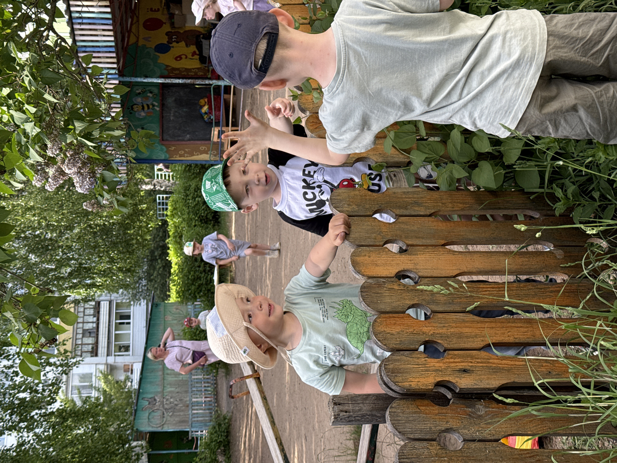 The image depicts three young children standing near a playground structure in a park. The playground has a wooden fence with decorative holes. One child wears a beige sun hat and a light green shirt with a dinosaur image. Another child wears a green bandana hat and a black and white shirt with a Mickey Mouse design, holding up his hand. A third child wears a dark blue baseball cap and a light gray shirt, standing behind the second child. In the background, two adults are walking through the park, and an apartment building is visible. The park appears to be in a residential area. The location is Chaikovsky, Russia.