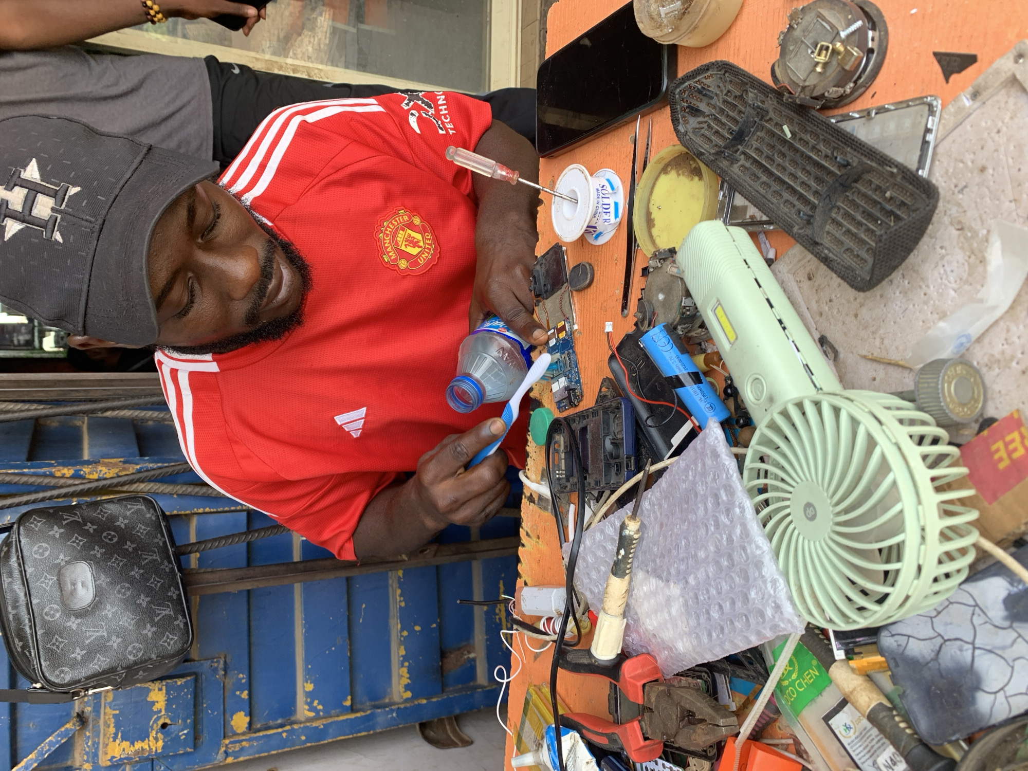 A man wearing a Manchester United jersey and a black Houston Astros baseball cap is seated at a table cluttered with electronics and repair tools. He is cleaning a circuit board with a toothbrush and a bottle of water. The table also contains components like a small fan, a car air vent, a roll of solder, and various circuit boards. A Louis Vuitton bag rests on a blue metal gate or railing to his left. The location is identified as Rafin Par, Nigeria based on available evidence within the image.