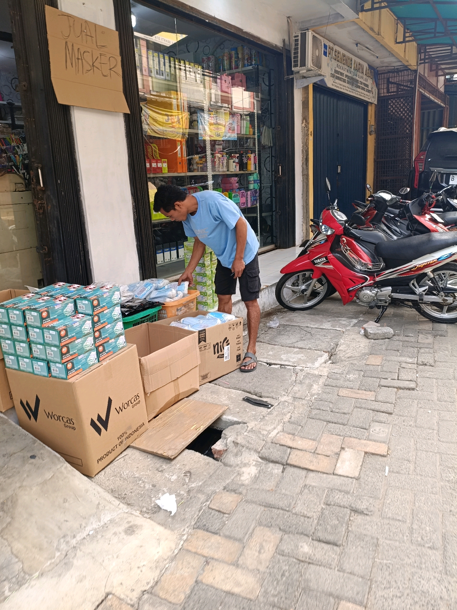 A man in a light blue t-shirt and black shorts is looking down at boxes of face masks outside a shop in Tangerang, Indonesia. The shop appears to be a small business selling various goods, as indicated by the shelves stocked with items visible through the glass windows. A sign on a piece of cardboard taped to the metal gate reads "JUAL MASKER" (Selling Masks). In front of the shop, several cardboard boxes and plastic crates filled with face masks are stacked on the sidewalk. One large cardboard box in the foreground has "Worcas Group" printed on it. A red and silver motorcycle is parked near the shop entrance, along with other motorcycles in the background. The street is paved with cobblestones and there is a hole in the pavement near the boxes. The overall scene suggests a commercial area with an emphasis on the sale of personal protective equipment.