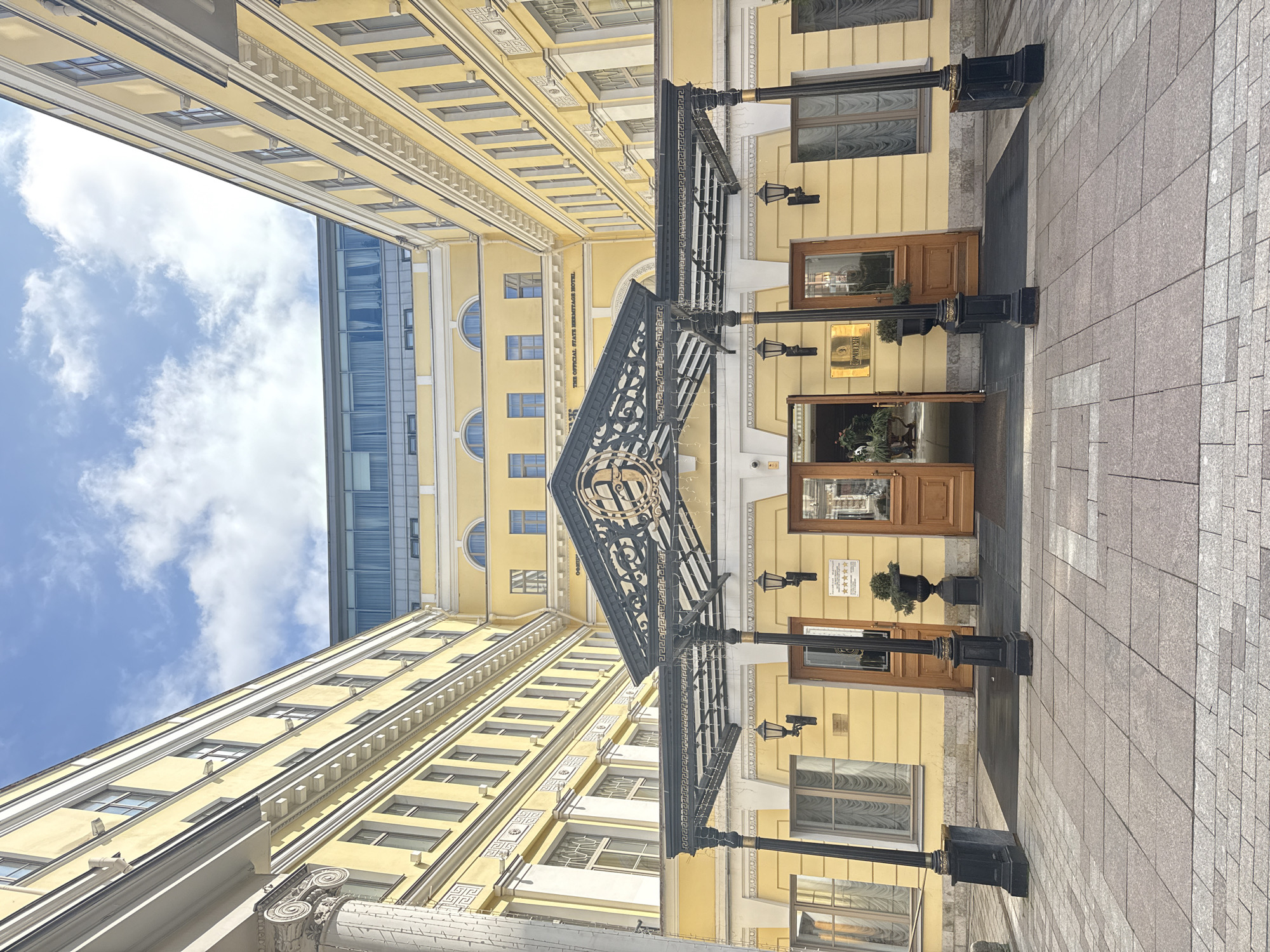 The image showcases the State Hermitage Museum Official Hotel in Saint Petersburg, Russia, viewed from a low angle looking up towards the building's entrance. The hotel is characterized by its yellow facade, multiple windows, and a decorative black canopy over the entrance. The entrance features wooden doors and small potted plants on either side. Adjacent to the hotel is a modern building with a glass exterior visible in the background. The sky is blue with scattered clouds, and the foreground depicts a paved walkway leading to the entrance.