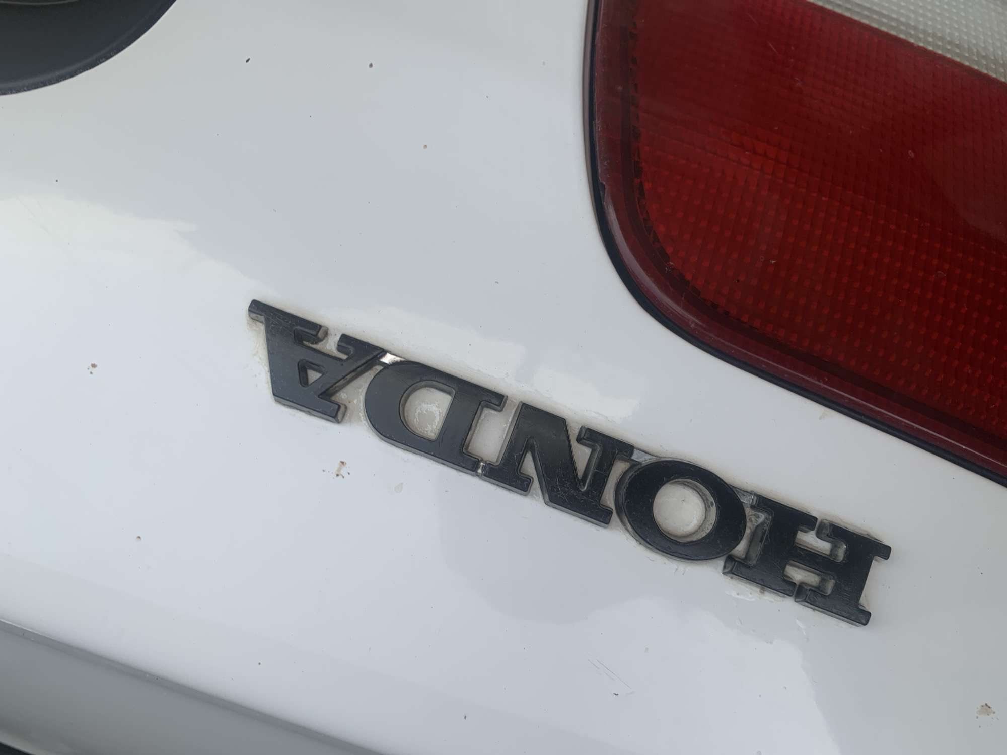 A black Honda emblem is mounted on the white exterior of a car, positioned to the right of the car's red taillight. The car appears to be outdoors, possibly in Jauro Yinu, Nigeria, given the context provided, although no specific city identifiers are visible within the image itself. The surface of the car exhibits minor dust and scratches, suggesting it may be in an area with dust or a slightly aged vehicle. The emblem's letters are raised and three-dimensional, with the 'H' being the leftmost character visible, followed by 'O', 'N', 'D', and 'A'. The overall scene is a close-up shot focusing on the car's branding.
