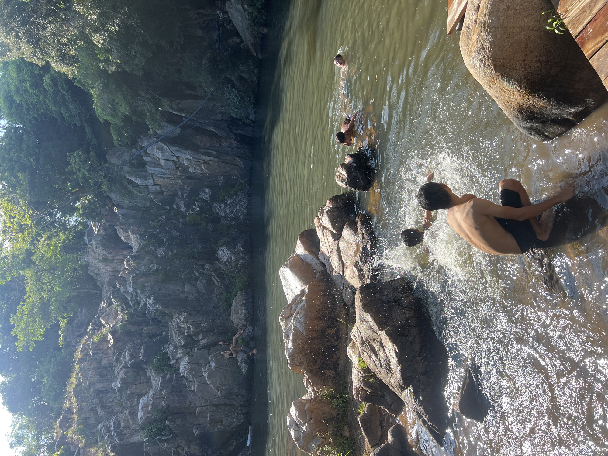 The image depicts a natural swimming area amidst a rocky landscape with lush vegetation. In the foreground, a young person is partially submerged in the water, appearing to crouch or sit near the edge. To their right, a large, smooth boulder sits on wooden planks. The water is disturbed by splashes, indicating movement and activity. In the middle ground, several other individuals are swimming or playing in the water. Further back, a rocky cliff face rises, with two people seated on rocks near its base. The water is a murky green, reflecting the surrounding trees and rocky formations. The scene is bathed in sunlight, with rays of light filtering through the trees, creating a bright and airy atmosphere. The location is identified as Vĩnh Hải, Vietnam.