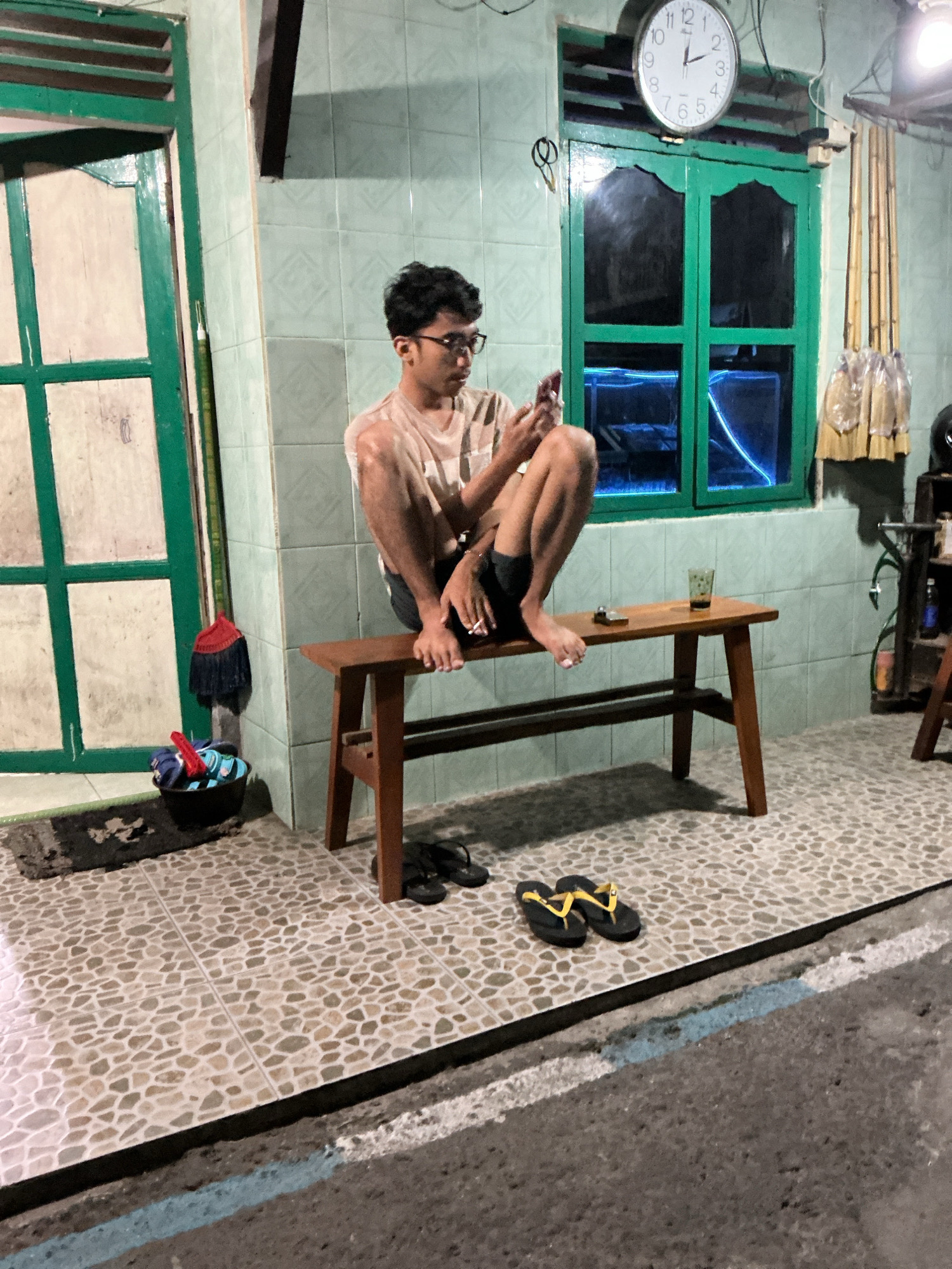 A young man with glasses and dark curly hair is sitting on a wooden bench in what appears to be an outdoor covered area, possibly a front porch or patio, in Magelang, Indonesia. He is wearing a light-colored t-shirt and dark shorts. He is holding a mobile phone in his hands and looking down at it. His left leg is crossed over his right, and his feet are bare. A cigarette is visible in his right hand.
The bench is made of wood and has two slatted supports. On the bench next to the man, there is a small glass with a drink and a metal object. In front of the bench, on the tiled floor, are two pairs of flip-flops, one black and one black with yellow straps.
The background features a wall tiled with light blue square tiles. To the left of the man is a green-painted door with glass panes. To the right of the man is a green-painted window with multiple panes, some of which show a blue light from inside. Above the window, a round analog clock with a white face is mounted on the wall. Further to the right, there is a collection of bamboo poles and a shelf unit with various items.
The floor is tiled with a mosaic pattern of irregular brown and beige stones. The area is illuminated by artificial light, creating shadows. A small bucket with some cleaning supplies, including a brush and a cloth, is placed near the door. A broom with a red and black head is leaning against the wall next to the door. The ground outside the tiled area appears to be a darker, rougher surface, possibly a road or pathway, with some white and blue markings. The overall impression is a casual, everyday scene in a residential setting in Magelang.