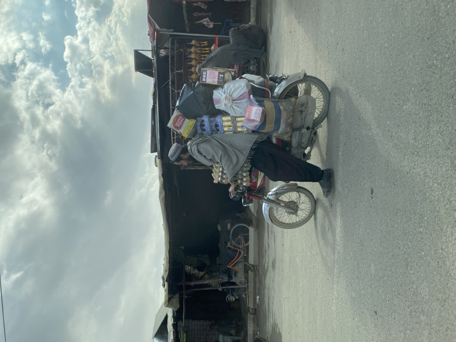 A bearded man, wearing a skullcap, a patterned shawl, and dark trousers, operates a red motorcycle heavily laden with cargo on an asphalt road in Kandahar, Afghanistan. He is positioned with his left foot on the ground, indicating a stop or slow movement. The motorcycle's front carries multiple stacked trays of eggs. The rear carrier is secured with a large black plastic bag, stacked items resembling beverage cans, a yellow box marked "SELERA PEDAS," several white cloth bags, a striped woven bag, and a smaller red-wrapped package.
In the background, a row of rudimentary market stalls or shops with corrugated metal and tarp roofs are visible. Within these stalls, a bicycle with an orange frame, hanging plucked poultry, and other meat products are discernible. A dark-clad figure is present within one of the stalls. A solar panel is mounted on a stall roof on the right. Another person is seated under a stall canopy on the far left. The sky is overcast with a mix of dark and lighter clouds.