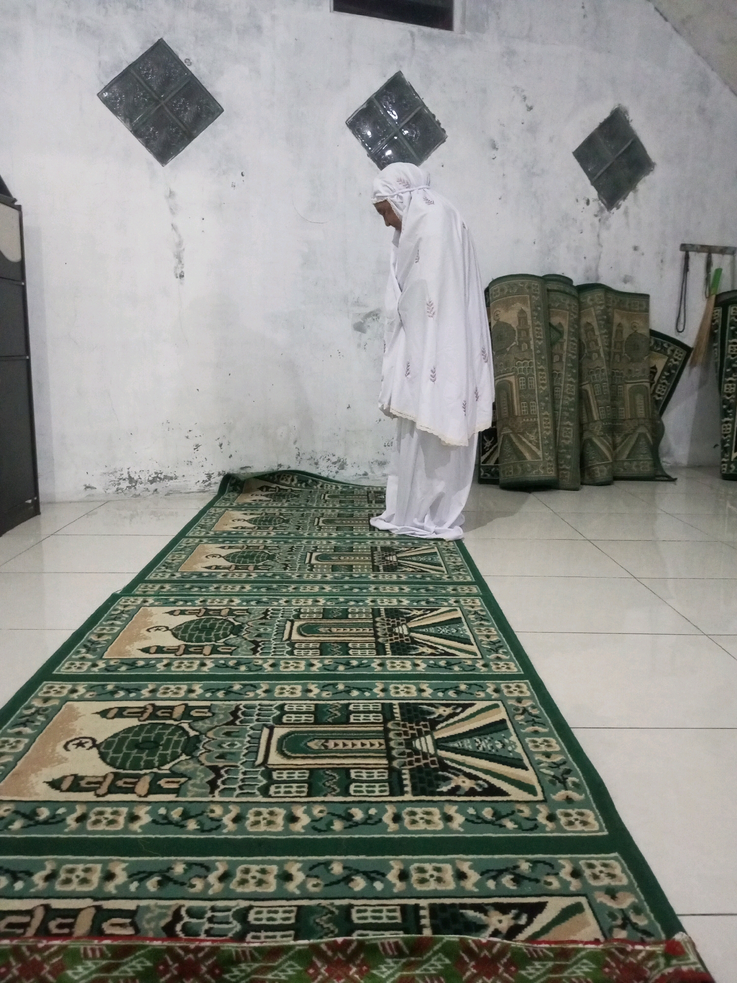 A person wearing a white hijab and white clothing is performing prayer on a prayer rug. The rug is green and cream with intricate patterns. Several other prayer rugs are stacked vertically against a wall in the background. The wall is white and textured, with three diamond-shaped decorative elements. The floor is tiled with white squares. The scene appears to be indoors, possibly in a room in Andongsili, Indonesia, used for prayer.