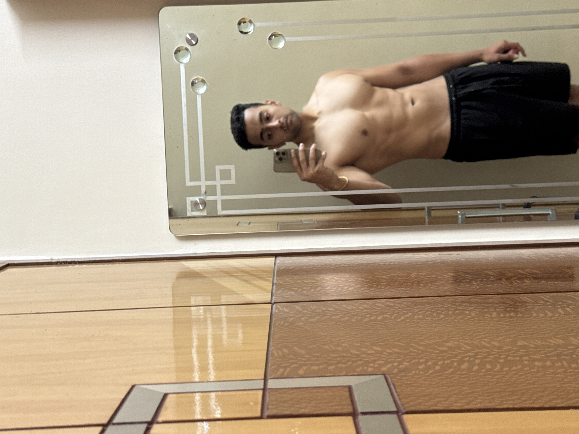 A man is taking a selfie in a mirror. He is wearing black shorts and no shirt. The mirror has a decorative frame with small, round, reflective embellishments. The background behind the mirror is a plain, off-white wall. Below the mirror, there is a wooden surface with a geometric pattern.
Without any further information, it is impossible to determine if the location is in Kolkata, India.