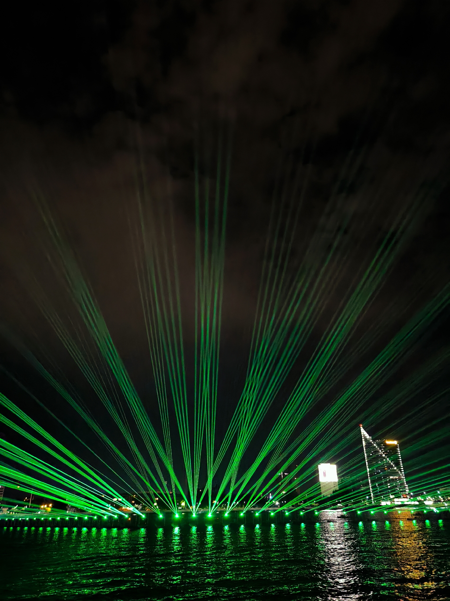 The image depicts a night scene in Riga, Latvia, with a panoramic view of a riverfront illuminated by a spectacular green laser light show. The lasers originate from the left and right sides of the frame, fanning out upwards and converging in the upper sky, creating a dramatic visual effect against the dark, cloudy night sky. The beams of light are sharply defined and extend outwards, some reaching high into the atmosphere.
Along the riverbank, a cityscape is visible, featuring modern buildings, including a tall, illuminated skyscraper with a distinctive pointed top on the right side of the image. Another large, rectangular illuminated screen or building stands to the left of the skyscraper. The lights from the city and the laser show are reflected on the rippling surface of the dark water in the foreground, creating a shimmering, green and yellow glow. The foreground water appears disturbed, suggesting movement from a boat or wind. The overall scene conveys a festive and technologically advanced atmosphere, characteristic of a large-scale public event or celebration in a modern urban setting. No people are clearly discernible in the image due to the distance and the nature of the light show.