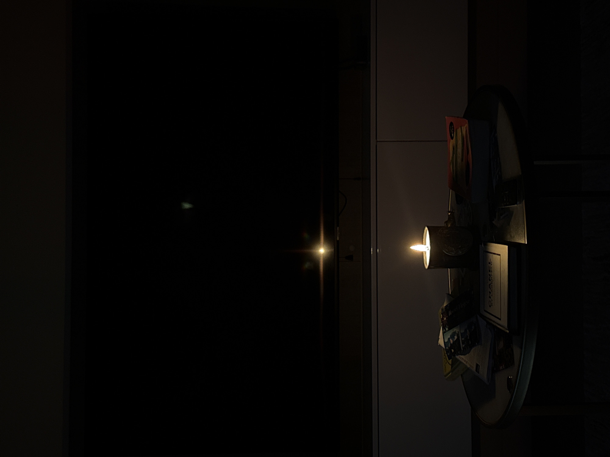 A dimly lit interior scene shows a dark gray refrigerator with various items magnetically attached to its surface. A small, cylindrical light fixture, likely attached to the fridge, illuminates the objects. Items include a CHANEL book, postcards, and what appears to be packaging for food or other goods. The lighting is focused on the fridge and the area immediately around it, with the rest of the scene in deep shadow. The reflection of the light source is visible on the dark surface to the left, indicating a smooth, reflective surface. No people are visible. No information is present to corroborate the location being Khimki, Russia.
