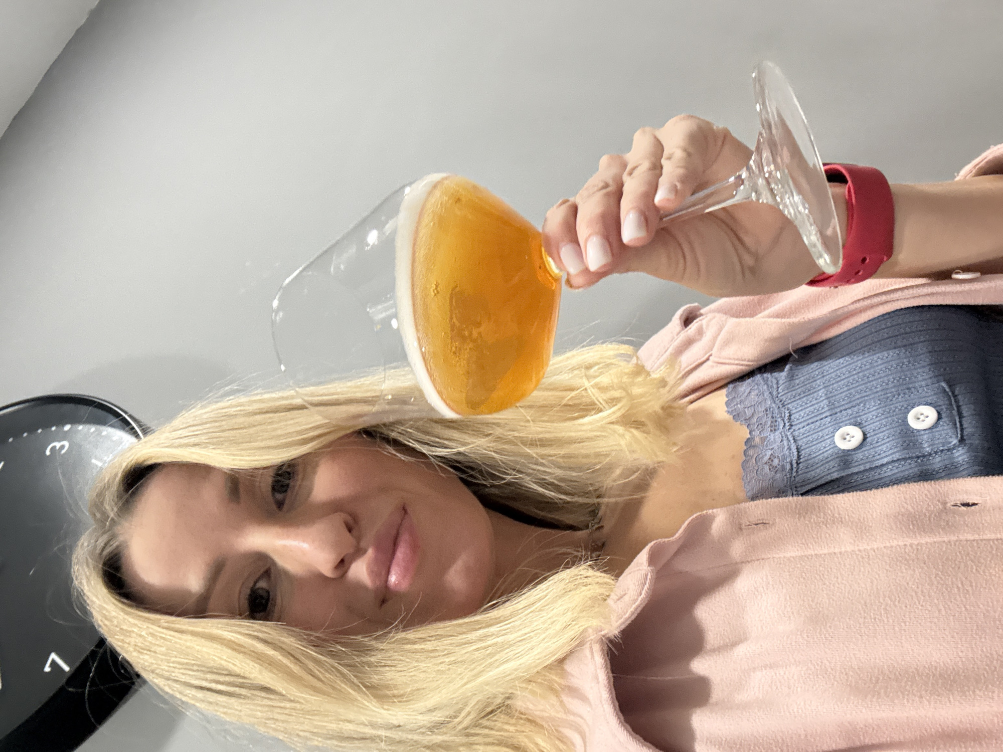 The image depicts a blonde woman holding a glass of beer, likely indoors, as suggested by the neutral-toned wall and partially visible clock. The woman is wearing a light pink button-down shirt over a blue lace-trimmed top with white buttons. She has a red smartwatch band on her wrist. The beer glass is tilted, with the amber-colored liquid and white head clearly visible. There is no explicit evidence within the image to confirm its location in València, Spain.