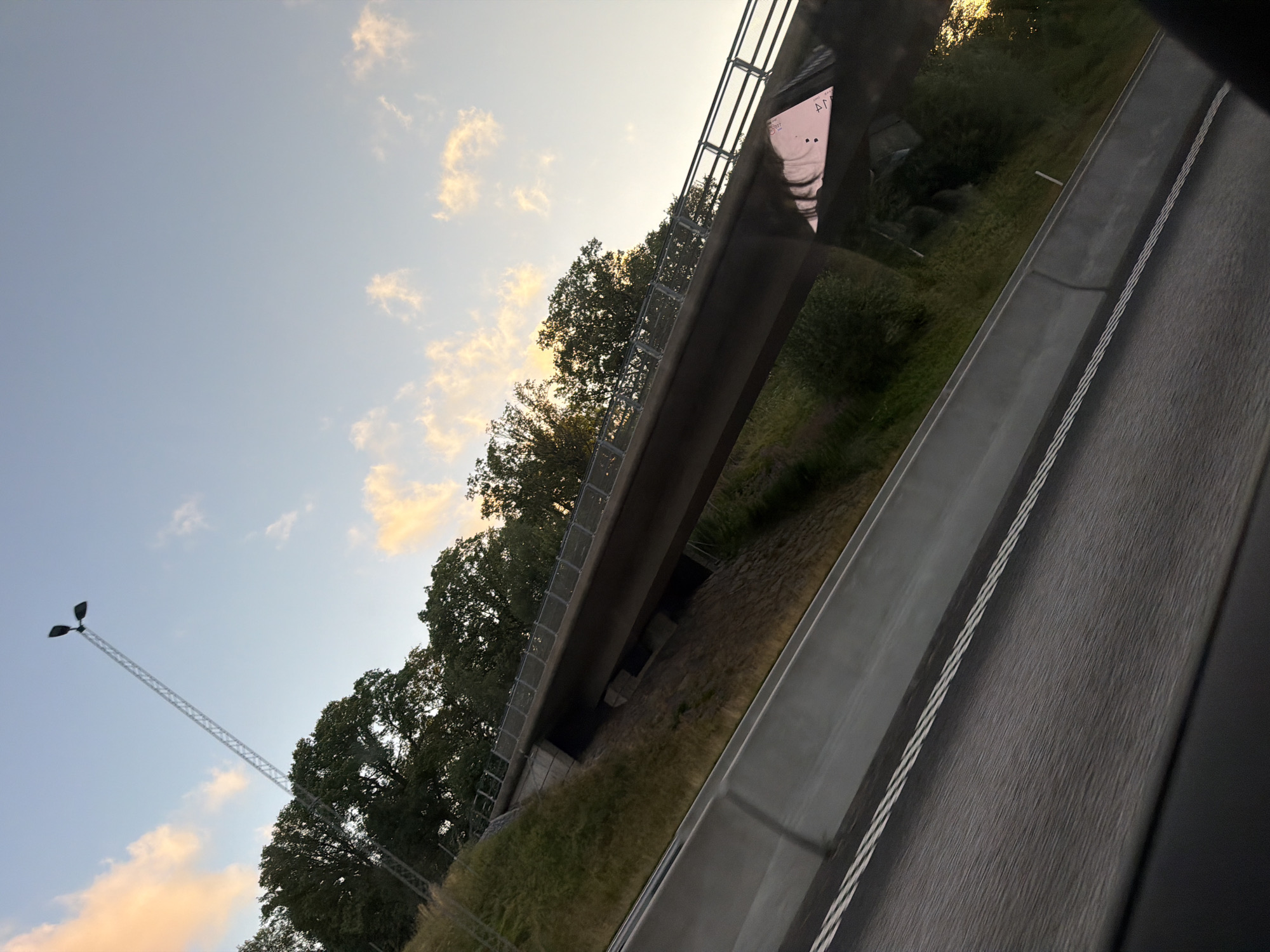 The image depicts an overpass on a highway in Munkedal, Sweden, during sunset. The sky is a pale blue with scattered clouds tinged with orange and pink from the setting sun. A tall, lattice-like lamppost with two lights stands prominently against the sky. Lush green trees line the embankment beneath the overpass, and the overpass itself has a metal railing. The highway, in the foreground, has a concrete barrier and double white lines painted on the asphalt, indicating a road in motion. The overall scene suggests a transitional moment of the day, with natural elements and infrastructure coexisting.
