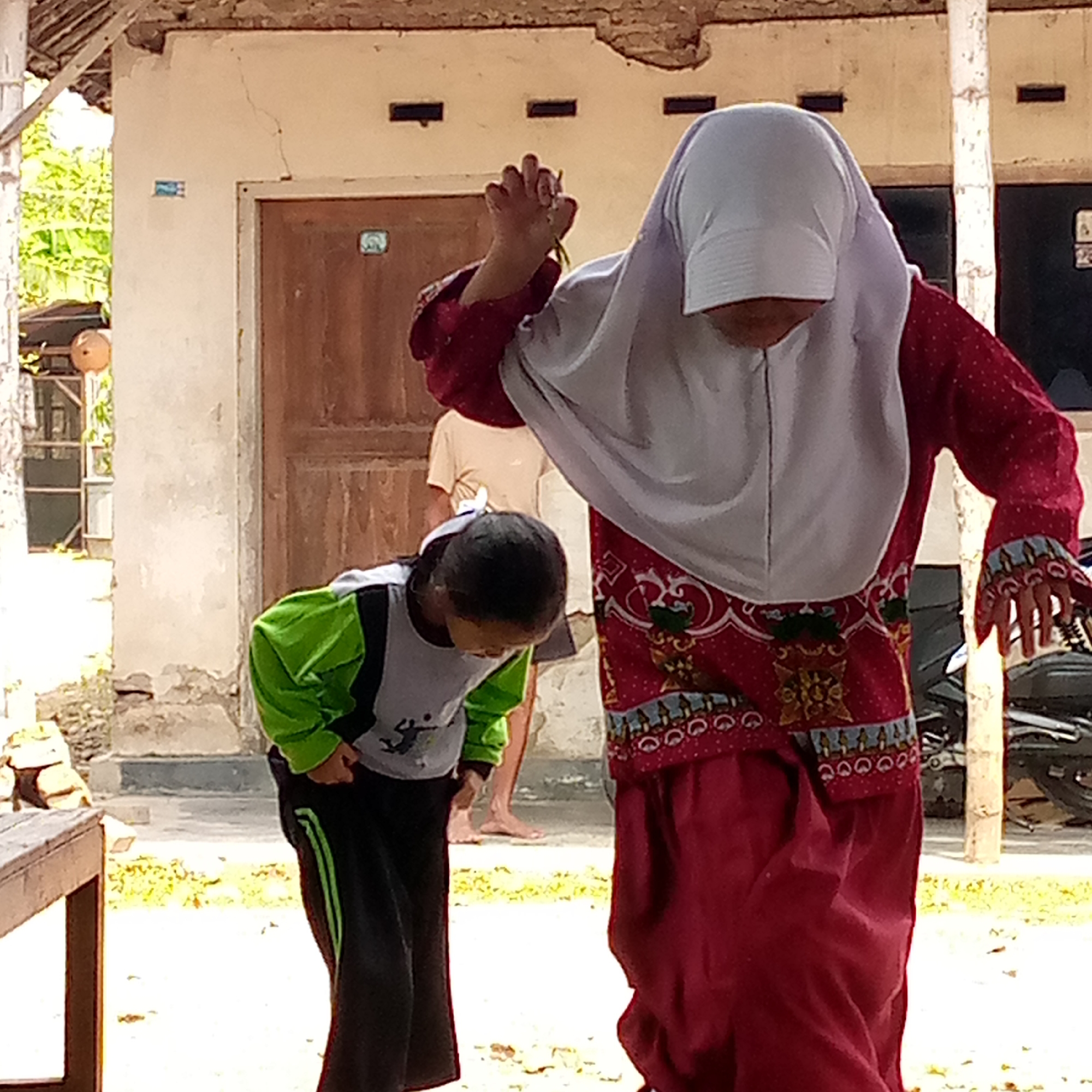 In Sumberwono, Indonesia, two individuals are pictured outdoors in front of a weathered building. The person on the right, wearing a red garment with white polka dots and a white hijab, is holding their hands up, with fingers extended. Their face is obscured by the hijab. To the left, a second person, appearing to be a child, is bent over with their head down, wearing a green and gray long-sleeved shirt and dark pants with a green stripe. Their dark hair is visible, and they are looking downwards. A third person, a young male wearing a light-colored t-shirt, is partially visible in the background between the two foreground figures. The building behind them has a rough-textured, light-colored exterior wall with a brown wooden door. Cracks are visible on the wall and the roof structure appears partially damaged. The ground is a light-colored concrete or dirt surface, with scattered yellow foliage. A wooden table or bench is in the foreground to the left, and a motorcycle is visible to the right. The overall lighting suggests a daytime scene with sunlight.