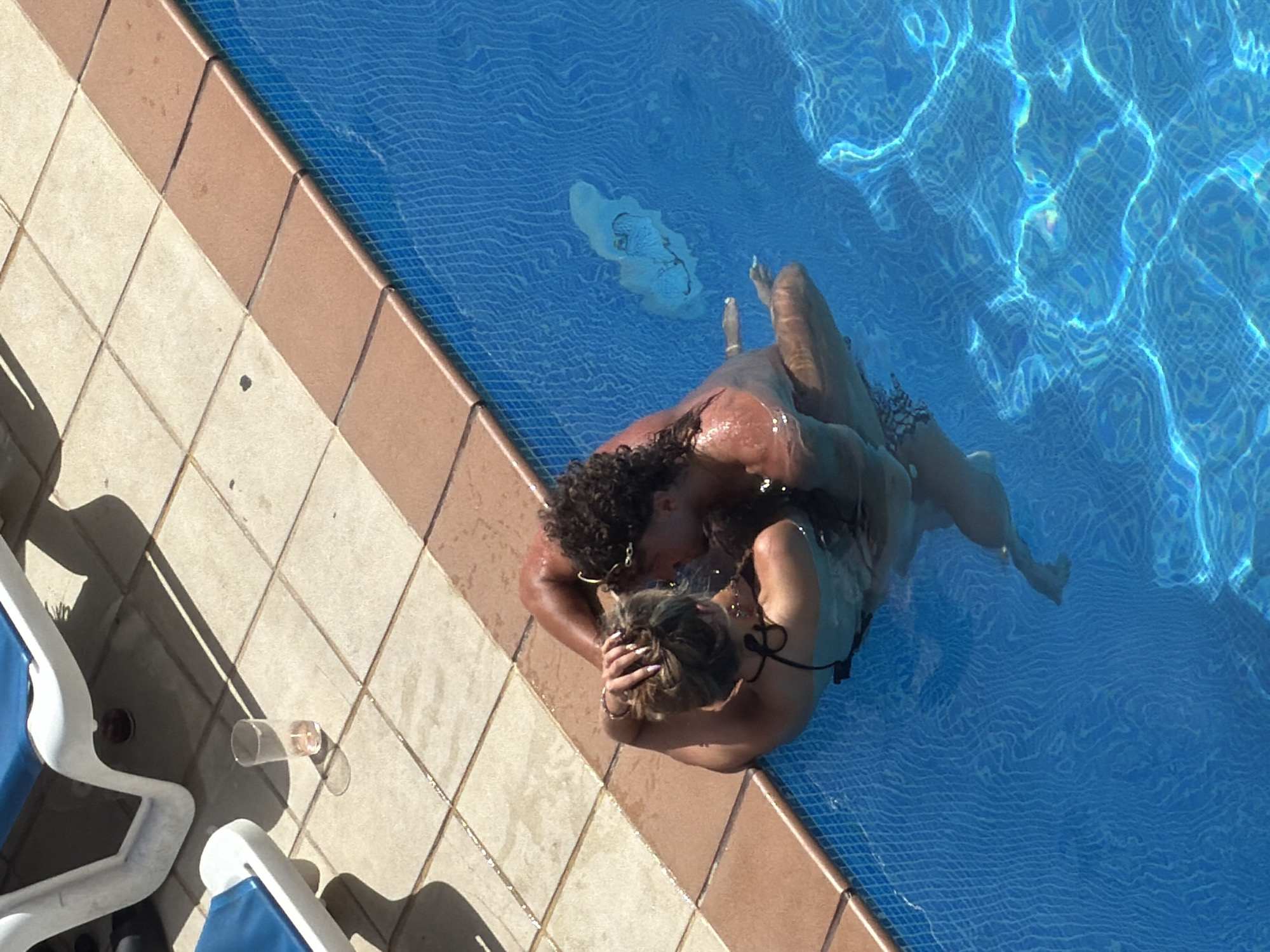 In Sant Antoni de Portmany, Spain, on a bright, sunny day, a couple is embracing in a swimming pool. The woman, with blonde hair, wears a black bikini and her arms are around the man's neck. The man, with curly dark hair, is shirtless and has his arms around the woman. They are submerged in the clear blue water. The poolside is tiled with beige and brown tiles, and a part of a white and blue lounge chair is visible on the left. A clear plastic cup containing a light pink liquid and ice is on the tiles near the lounge chair. The pool water exhibits a shimmering effect due to sunlight reflection.