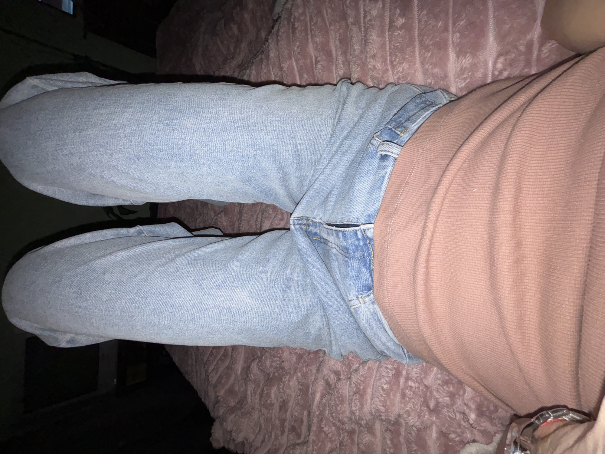 The image shows a person from the waist down, wearing light-wash denim jeans and a coral-colored ribbed top. The person is lying down on a plush, pink surface, possibly a bed or a couch. The background is dark and out of focus, with some indistinct shapes suggesting a room. There are no visible indications of the city or country.