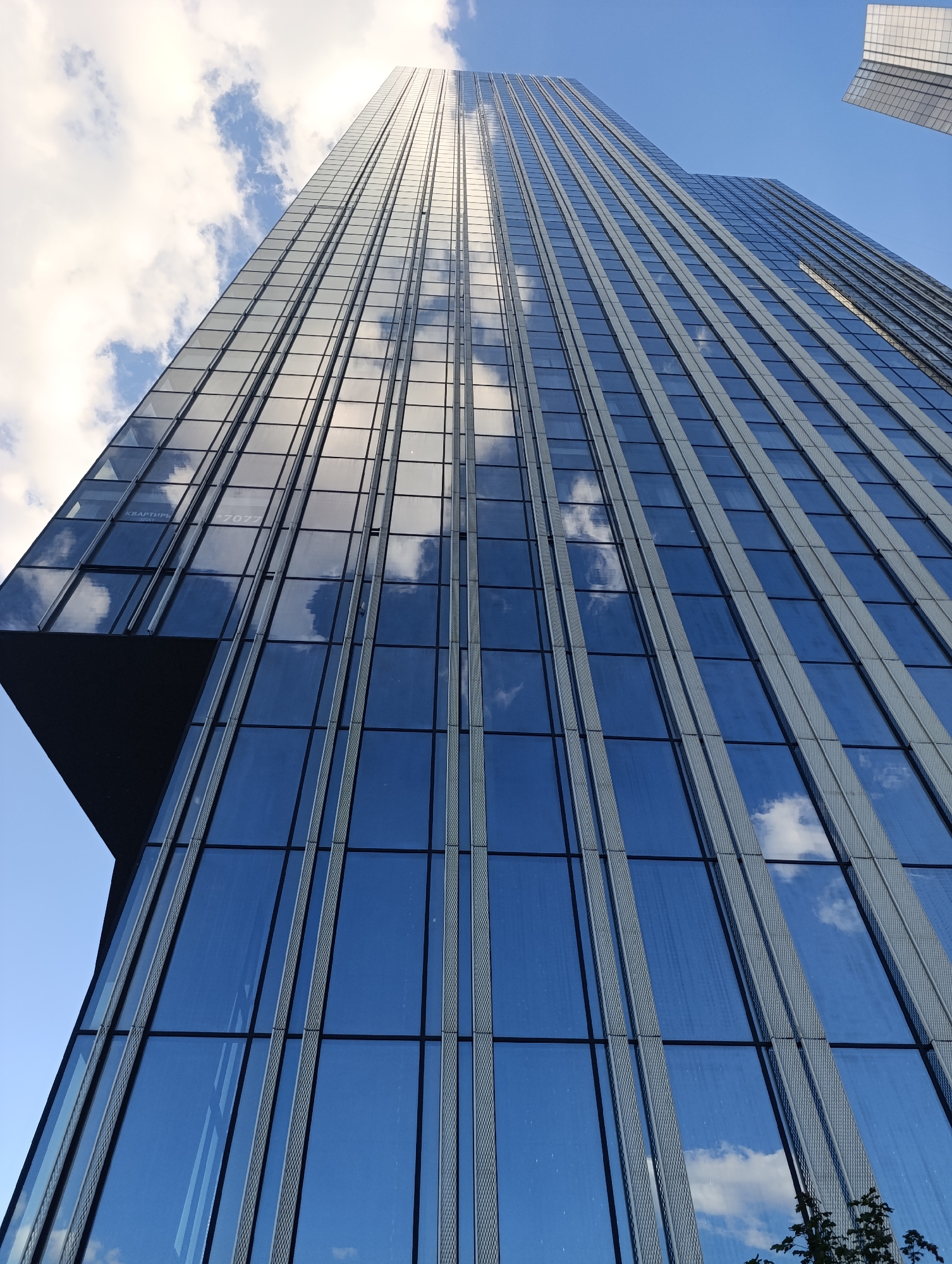 The image captures a striking low-angle view of a modern skyscraper in Moscow, Russia, showcasing its towering glass and steel architecture against a vibrant blue sky with scattered white clouds. The building's façade is predominantly made of reflective blue glass, which perfectly mirrors the clouds and sky, creating a dynamic interplay between the structure and its environment.
Vertical silver or gray-toned mullions, featuring a textured or patterned design, run up the entire height of the building, providing a strong linear element to the composition. The bottom left portion of the building reveals a distinct architectural feature, possibly a cantilevered section or a setback, which adds depth and complexity to its design. In the far top right, a sliver of another building is visible against the clear sky. A small amount of green foliage, likely from a tree or bush, can be seen in the bottom right corner of the frame.
The scene is illuminated by bright daylight, indicating clear and sunny weather. Visible on one of the glass panels in the lower left quadrant are Cyrillic characters that spell "КВАРТИРЫ" (Kvartiry), which translates to "Apartments," along with the numbers "7077." Above this, fainter text appears to include the word "GRAPHICS." The image emphasizes the scale and sleekness of the urban landscape.