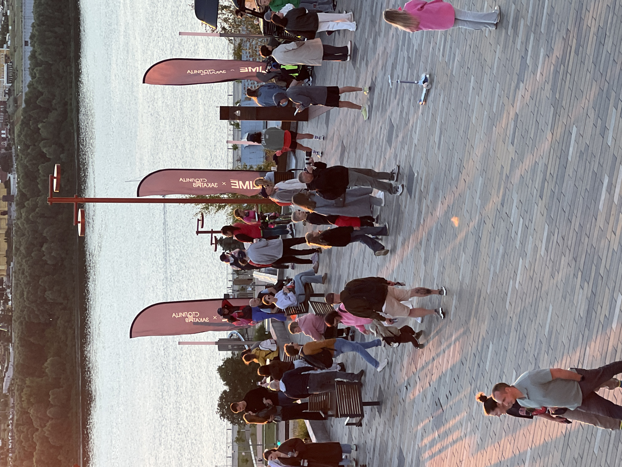 A group of people are gathered outdoors on a paved area next to a body of water. Many individuals are standing and appear to be participating in a yoga or exercise class, with some in various poses. Others are sitting on benches or standing around watching. The setting suggests a waterfront promenade, with trees and buildings visible in the background across the water. The sun is low in the sky, casting long shadows, indicating it is likely late afternoon or early evening. The weather appears to be pleasant. A few flags with text are visible, one prominently displaying "CLOUNIOT" and "TIME". A child's scooter is also visible on the ground. The overall mood seems relaxed and active.