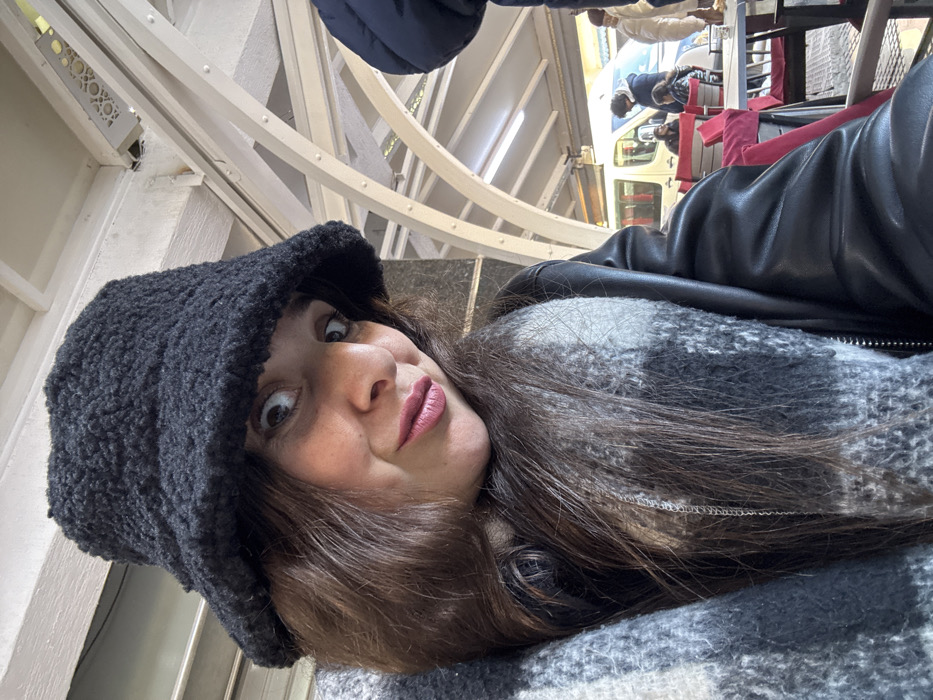 A female subject is depicted in a close-up, selfie-style photograph, positioned in the left foreground of a covered outdoor commercial area in Azrou, Morocco. She wears a black textured bucket hat, a black leather-like jacket, and a grey and white plaid scarf. Her dark hair is visible beneath the hat, and her facial expression shows wide eyes and slightly pursed lips, with pink lipstick. The background reveals light-colored arched structural beams and a flat ceiling, characteristic of a cafe terrace or similar public space. In the mid-ground, dark-topped tables and chairs with red cushions are arranged. Multiple individuals, primarily blurred, are visible in the background; one person in a dark blue puffer jacket is partially seen to the subject's right, and other patrons are seated at tables. A white passenger van is parked further back beyond the seating area.