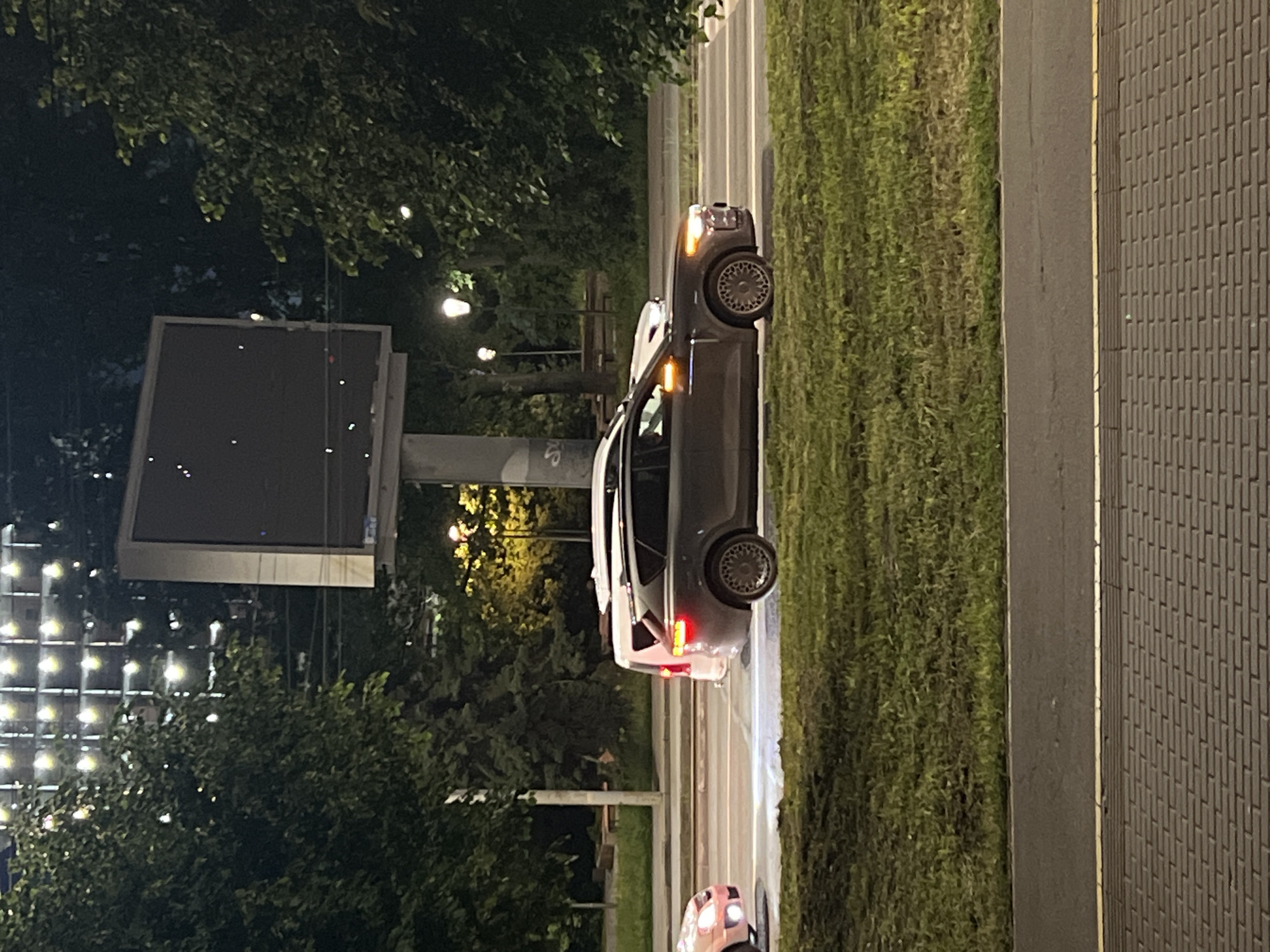 The image depicts a nighttime urban street scene in Izhevsk, Russia. A dark-colored car, possibly an SUV or crossover, is prominently featured on a paved road, positioned slightly to the right of the center. Its amber hazard lights or turn signals are flashing at the front and on the side mirror, and its red taillights are illuminated. The car is equipped with distinctive multi-spoke alloy wheels.
To the left of the road is a lush, grassy median strip, densely lined with trees whose dark foliage fills much of the left side of the frame. Beyond the trees, on the far left, a large, rectangular billboard mounted on a pole is visible, appearing dark and unlit. In the background on the left, a structure with numerous square-patterned lights casts a bright glow through the trees. Several warm-toned streetlights are also visible throughout the scene, illuminating parts of the road and trees.
To the immediate right of the paved road, there is a dark gray, textured surface, possibly a sidewalk or another part of the road infrastructure. The front lights of another vehicle are faintly visible in the bottom right corner of the frame. The overall atmosphere is that of a quiet, artificially lit night street. No discernible text is visible in the image.