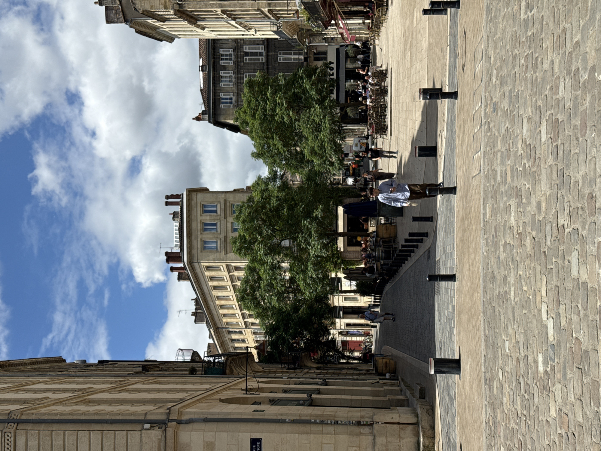 The image depicts a view down a European street, likely in Bordeaux, France, characterized by its Haussmannian-style architecture and cobblestone pavement. Tall, light-colored buildings with arched ground floors line the left side of the street. On the right, buildings with darker stone facades and numerous windows are visible, interspersed with outdoor seating areas for cafes and restaurants. Large, leafy trees are planted along the street, providing shade.
In the foreground, a man in a light blue shirt and brown pants walks away from the camera. Further down the street, several other individuals are present, some walking and others seated at outdoor tables. A few bicycles are also visible. The sky is bright blue with scattered white clouds, suggesting pleasant weather. The scene conveys a typical urban atmosphere, with people enjoying the outdoors and the architecture of the city.