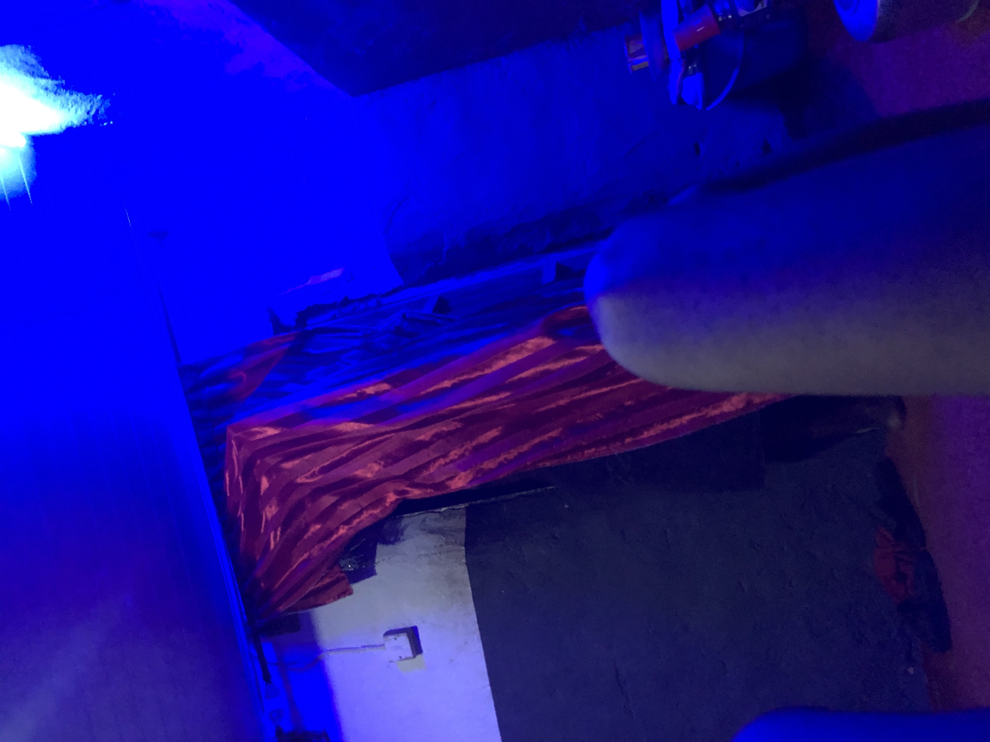 This image depicts an indoor scene characterized by dim, predominantly blue artificial lighting. The location is an unknown establishment within the city of Unknown, Unknown.
The scene features a prominent red, vertically hanging curtain or drape with visible folds, positioned slightly right of center. To the left of the curtain, a two-toned wall is visible: the upper section is light grey or off-white, and the lower section is dark grey or black, both heavily saturated by the blue light. A white electrical outlet is mounted on the upper wall section. In the upper left corner, a dark horizontal element, possibly a beam or conduit, is present.
The upper right portion of the image shows a strong, brighter light source, emitting a white-blue or cyan glow, partially illuminating the ceiling and an adjacent wall. The area to the right of the red curtain is largely in deep blue shadow, but some indistinct objects can be discerned in the lower right, including what appears to be parts of a shisha or hookah, with metallic and red components.
In the immediate foreground, a significant portion of the lower right is occupied by a blurred, elongated, light-toned object, consistent with a human limb (e.g., a knee or thigh), indicating the presence of a person, likely the photographer. The floor beneath the objects and limb appears dark and textured, exhibiting a reddish-purple hue in areas. No specific actions or interactions are depicted beyond the presence of objects in a static environment.